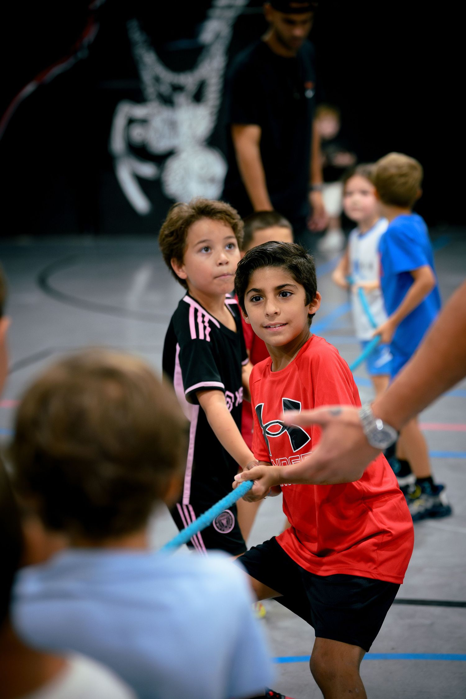 Children playing tug-of-war in a gym, with a boy in a red shirt leading the team during an indoor sports activity.