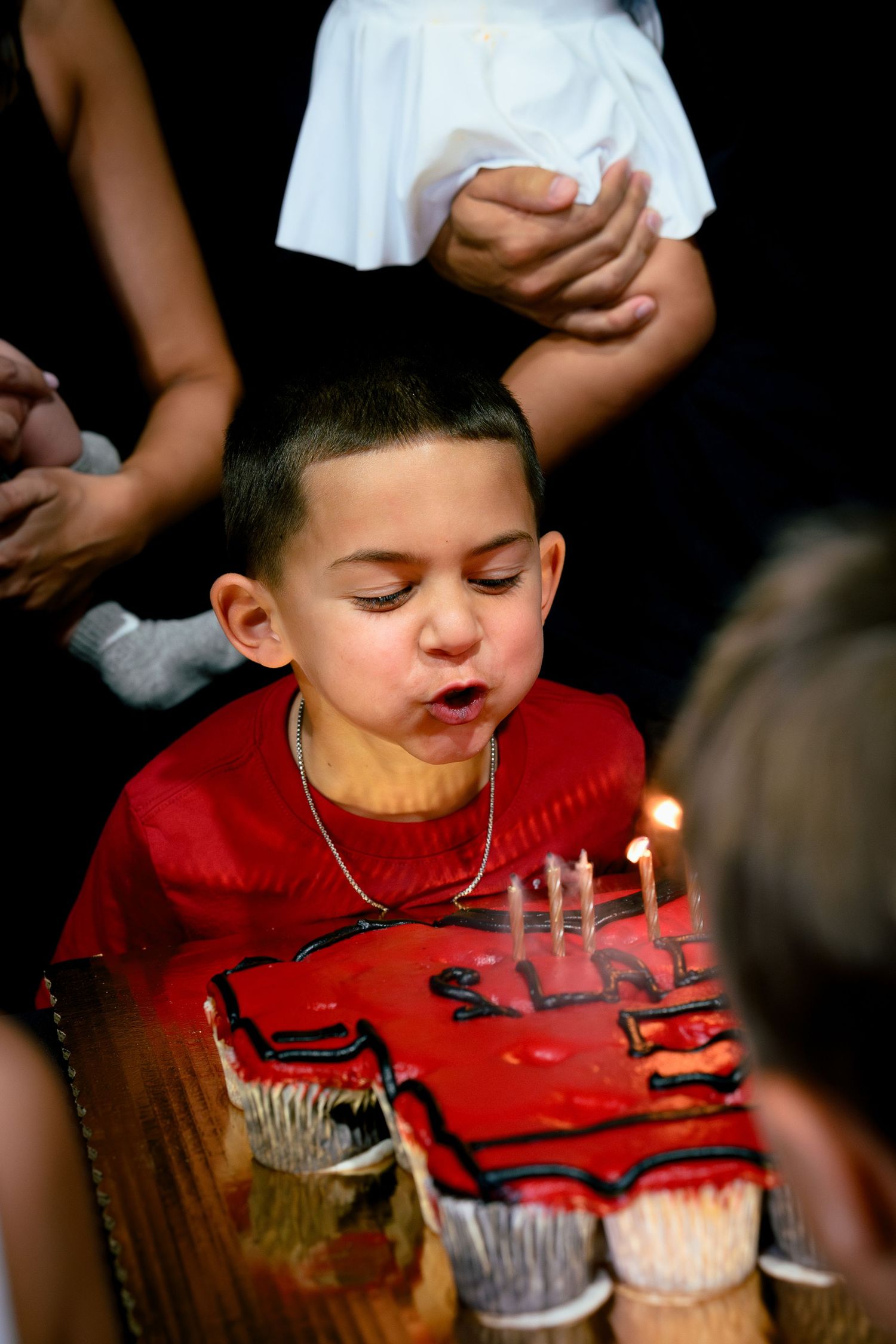 Young boy in red shirt blowing out birthday candles on a decorated cake surrounded by family.