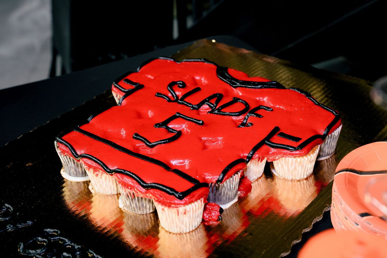Red jersey-shaped cupcake cake with black frosting reading SLADE 5 on a gold tray.