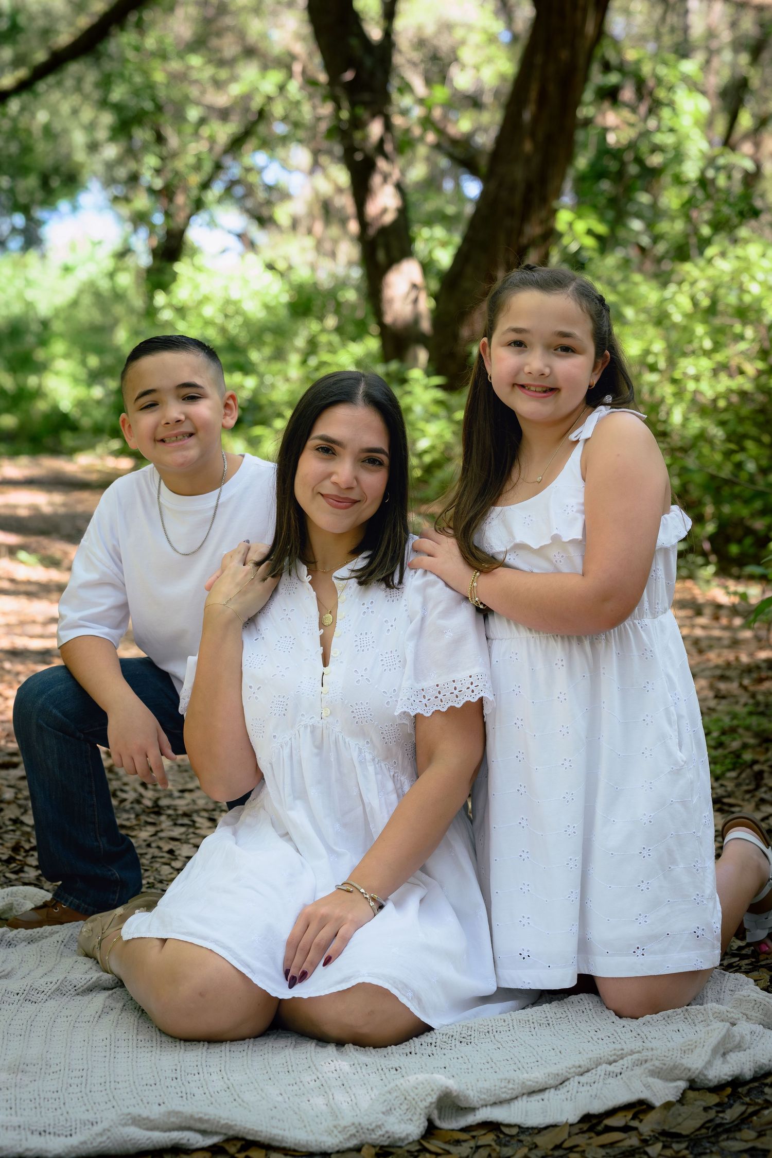 A woman and two children in white outfits pose together on a blanket in a lush green outdoor setting.