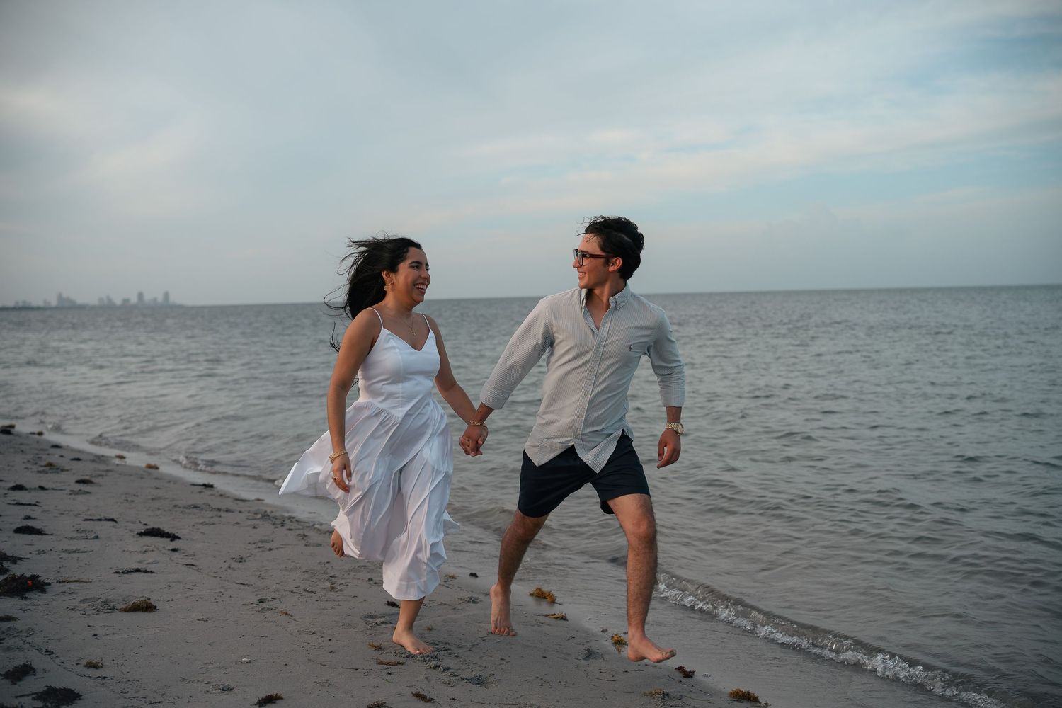 Couple holding hands and walking barefoot on a sandy beach by the ocean on a cloudy day.