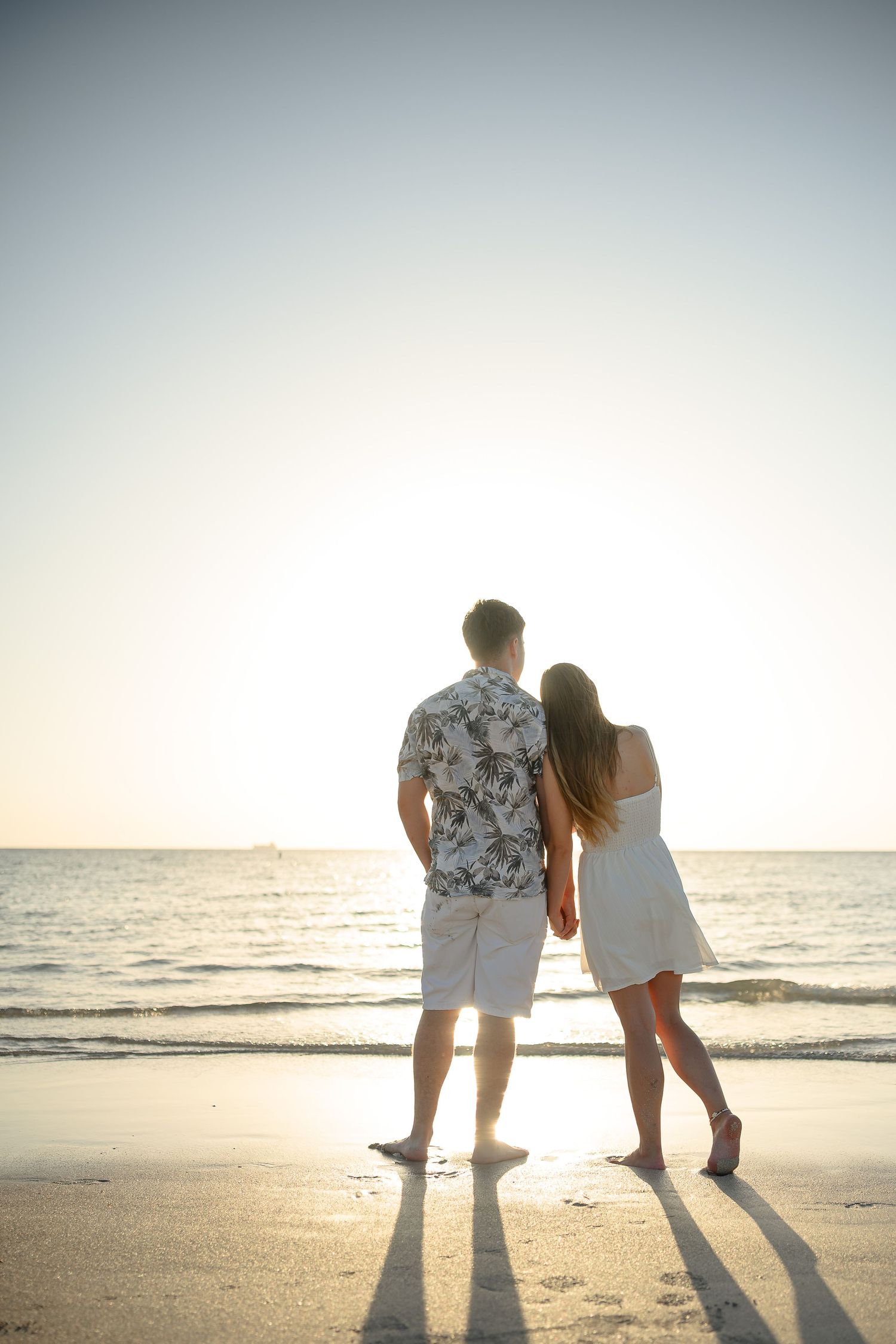 Couple holding hands on a sandy beach at sunset, facing the ocean, man in floral shirt and woman in white dress.