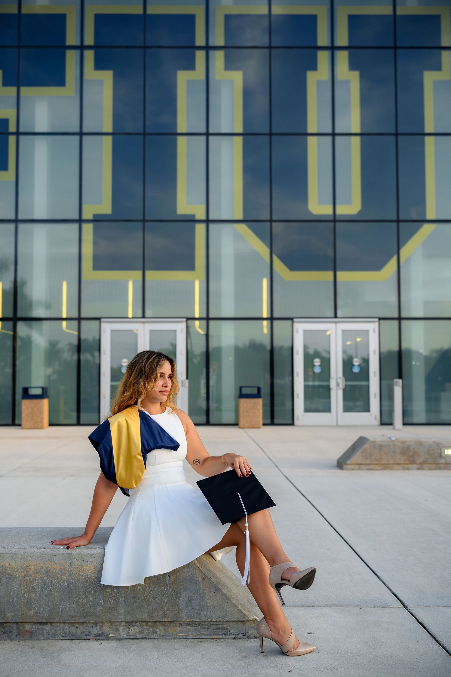 Woman in white dress holds graduation cap while sitting outside a modern university building.