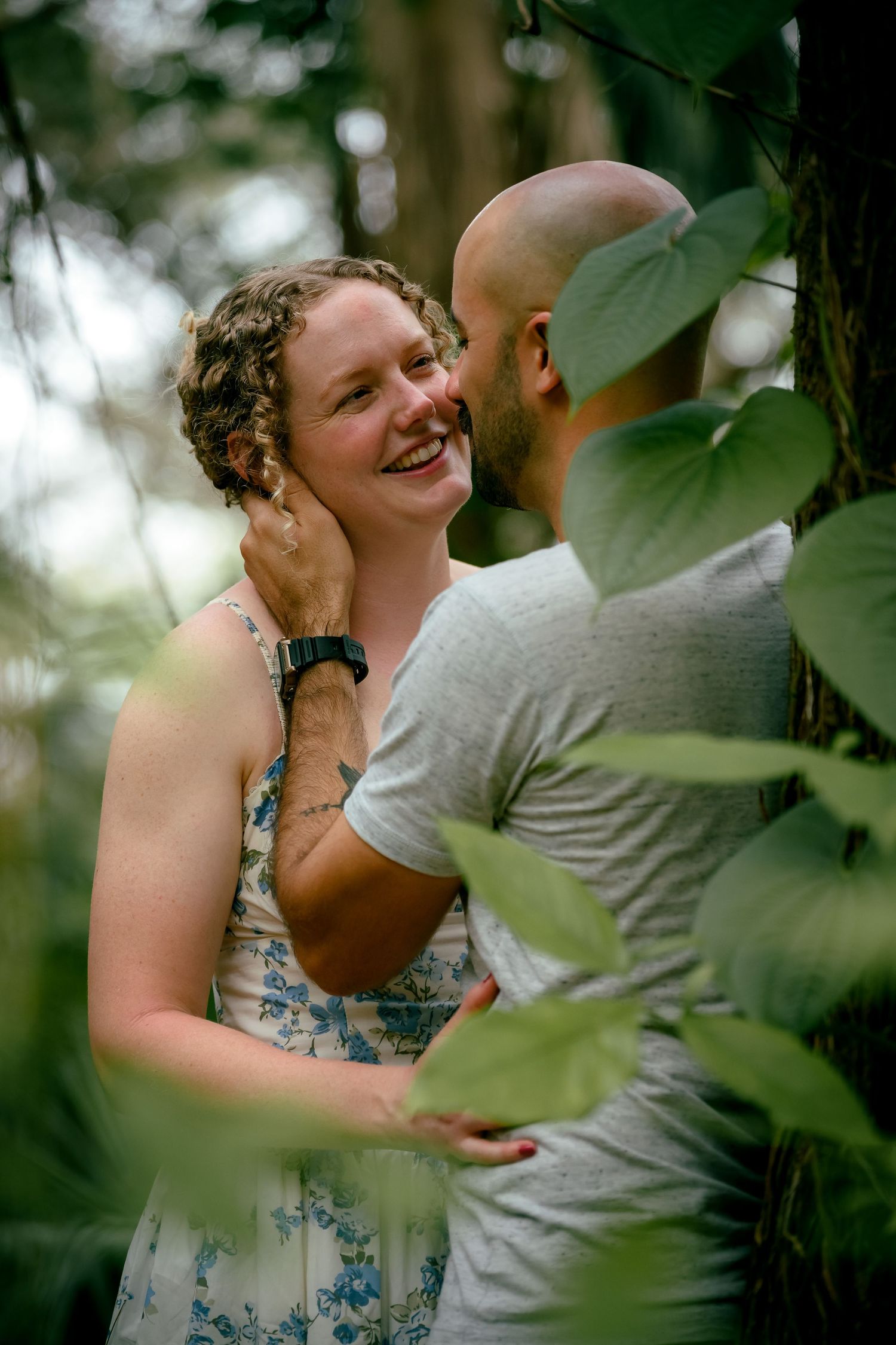 A smiling woman in a floral dress embraces a bald man among lush green leaves in a romantic outdoor forest setting.