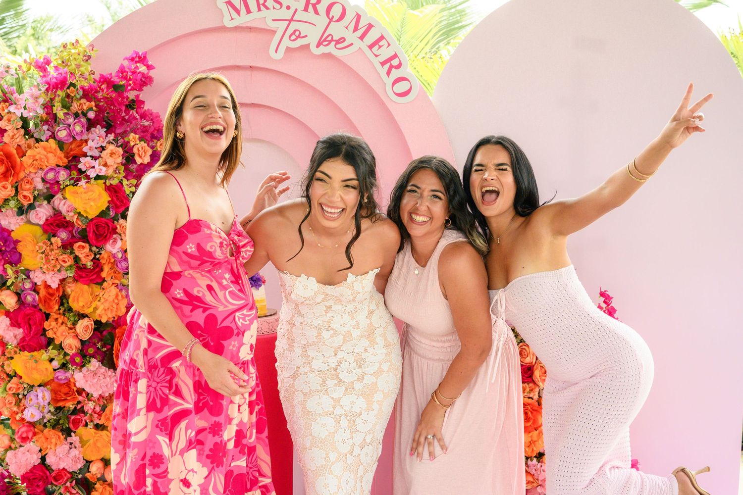 A group celebrates joyfully in front of a pink floral wall at what appears to be a bridal shower or wedding event.