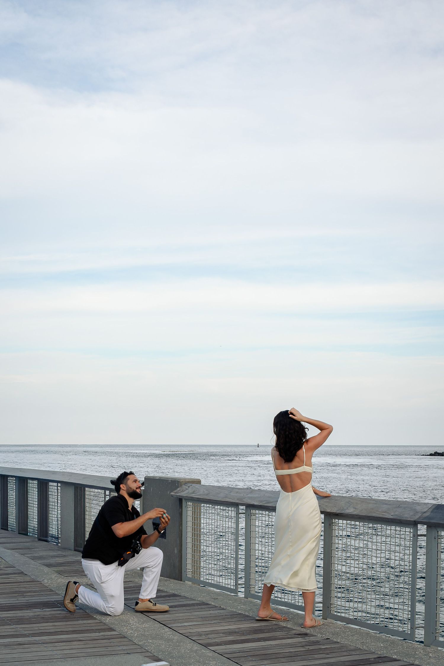 Man proposes on one knee to woman in white dress on a seaside pier with ocean views.
