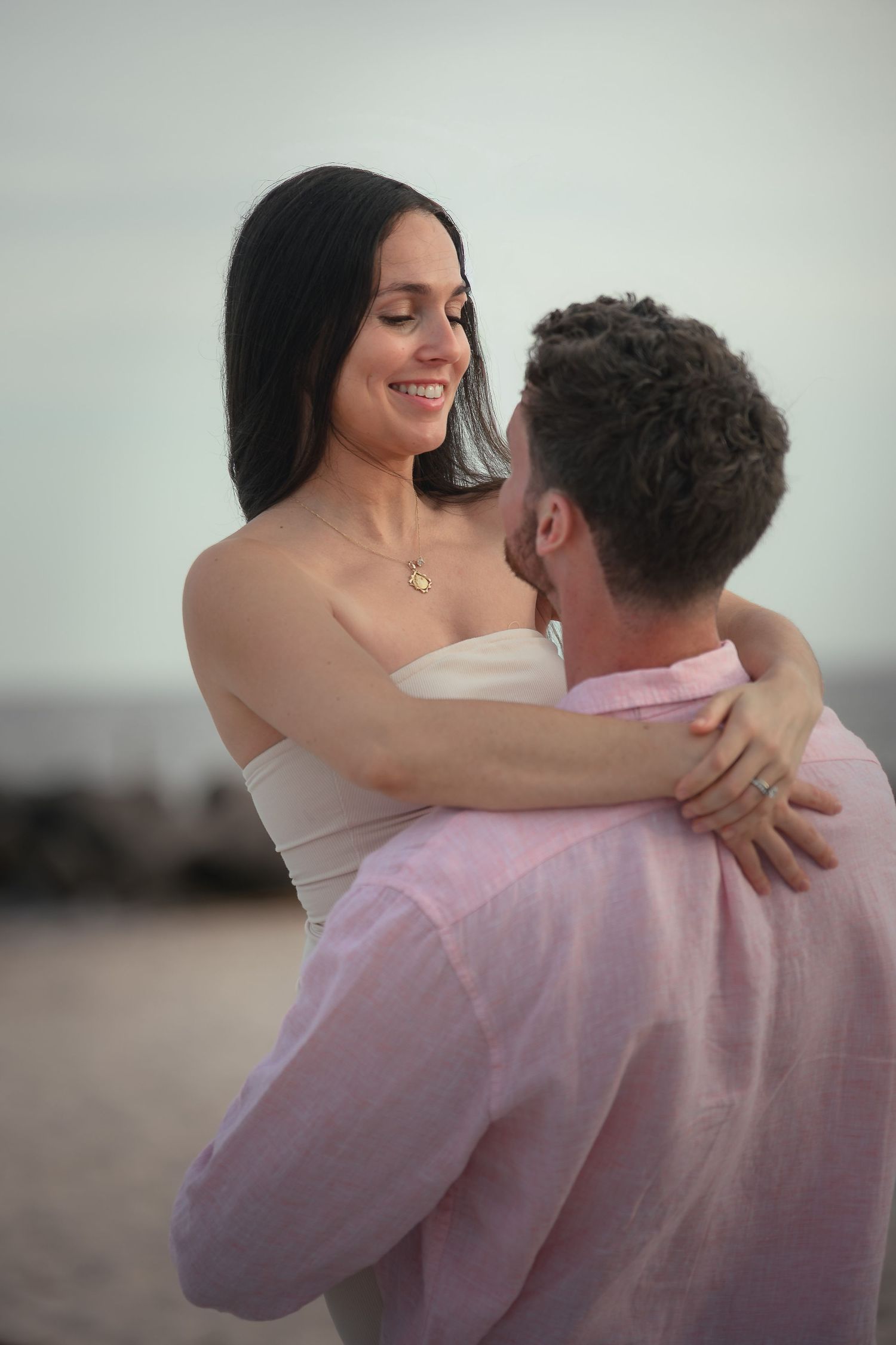 A couple embraces on a beach, woman in white top smiling at man in pink shirt, romantic outdoor portrait.