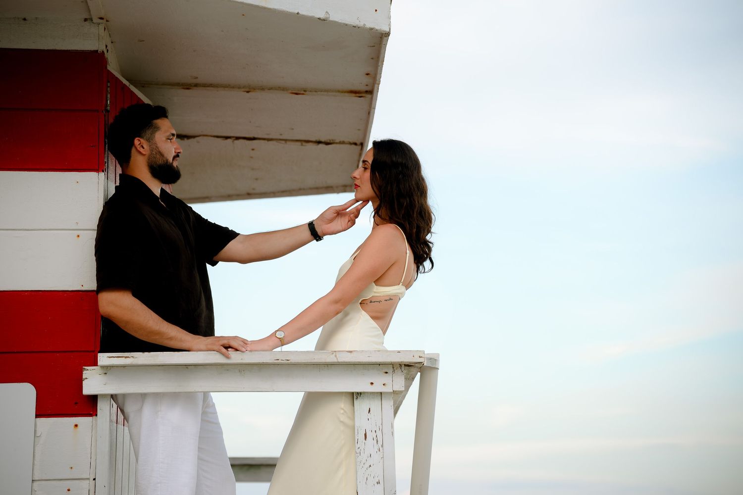 Couple sharing a romantic moment on a lifeguard stand, man tenderly touching womans face, ocean backdrop.