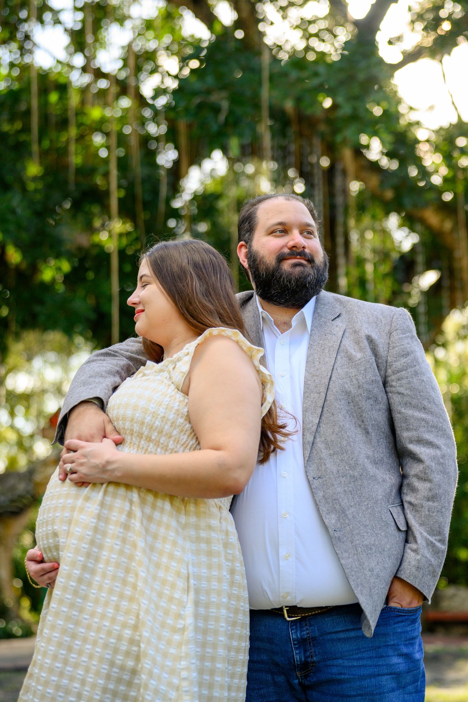 Pregnant woman in yellow dress embraced by bearded man in gray blazer, posing outdoors among lush green trees.