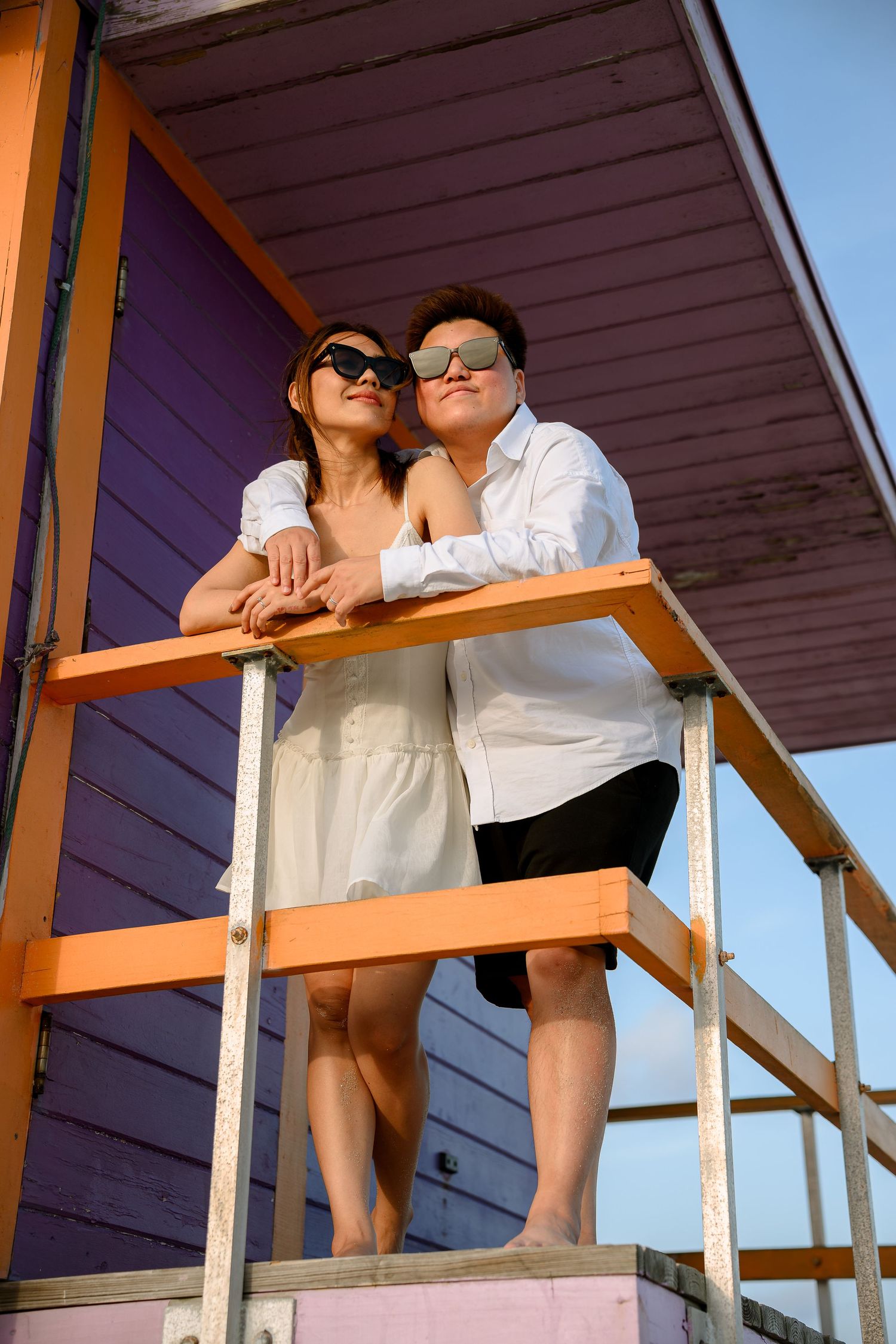 Couple in sunglasses leaning on railing of colorful purple and orange beach hut under blue sky.