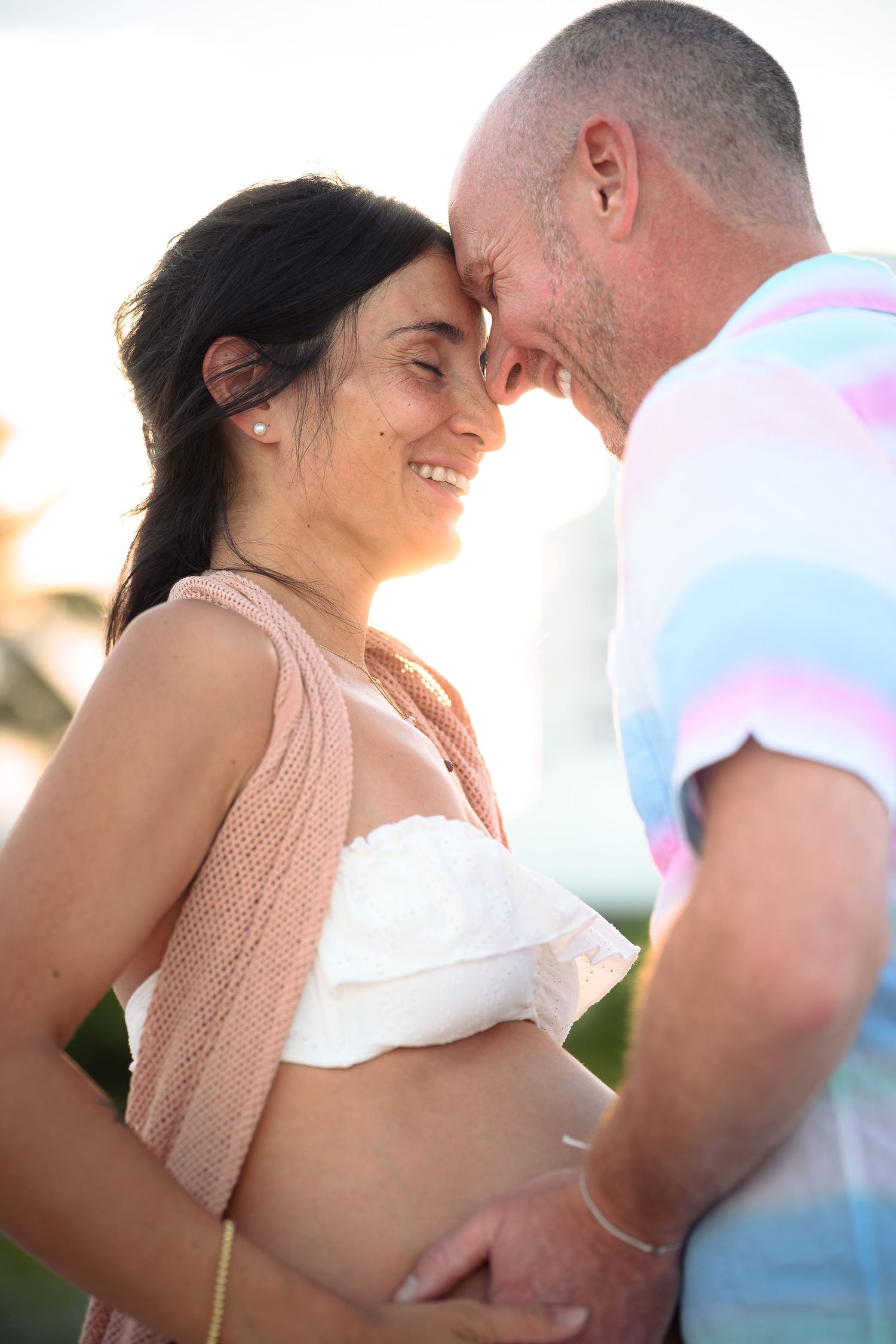 Happy pregnant couple sharing a tender forehead touch outdoors during golden hour, woman smiling in white crop top.