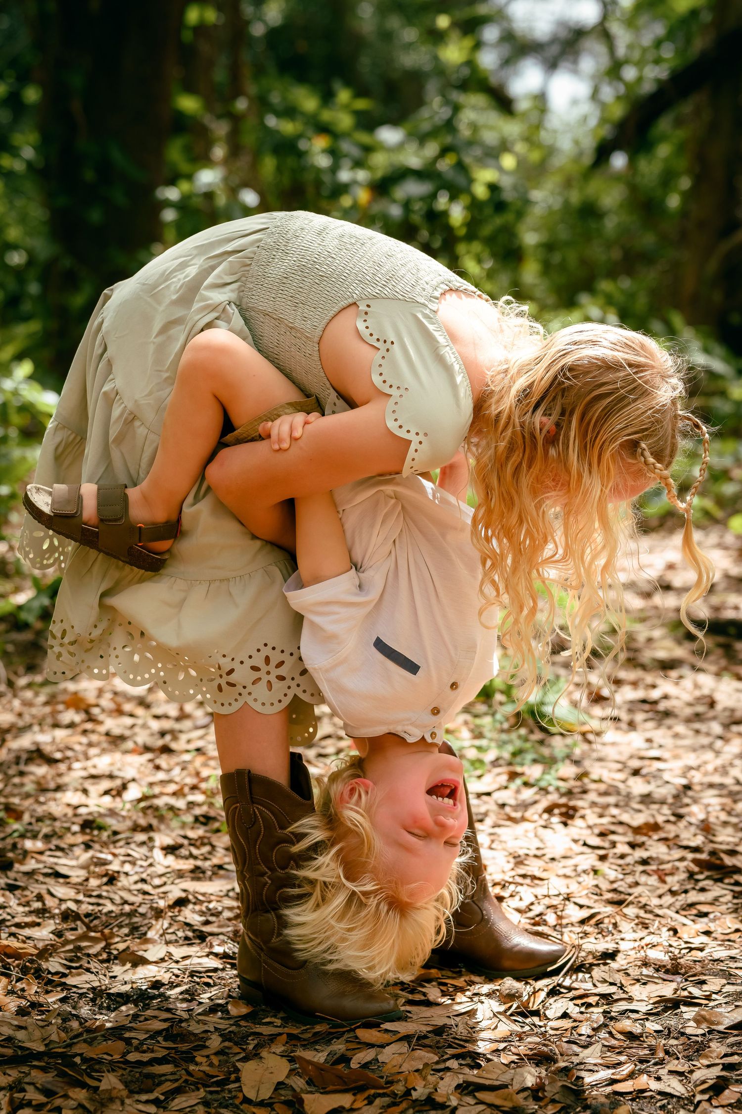 Girl in lace dress holds toddler upside down playfully in a sunlit forest with fallen leaves.