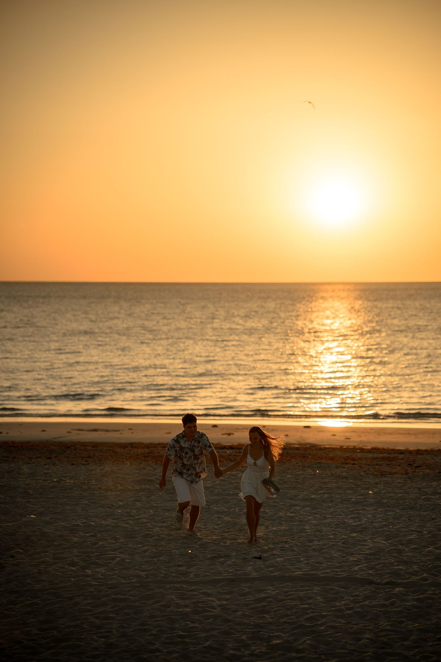 Couple holding hands running on a sandy beach at golden sunset with sun reflecting on calm ocean waves.