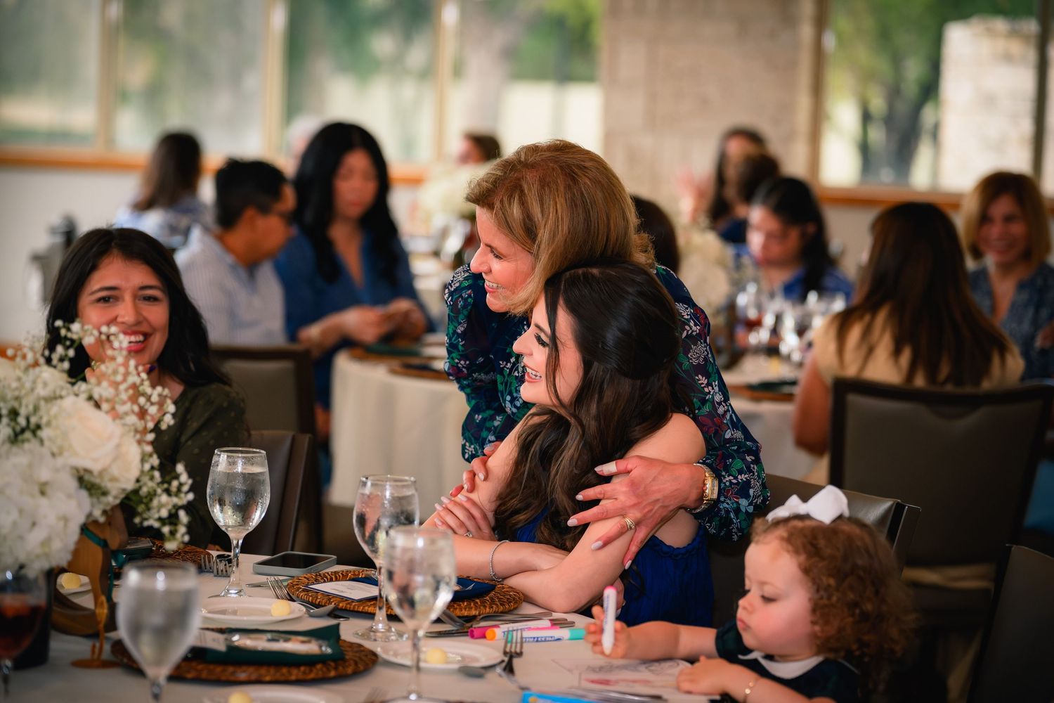 Guests sharing warm hugs and laughter at an elegant baby shower celebration in a bright venue.