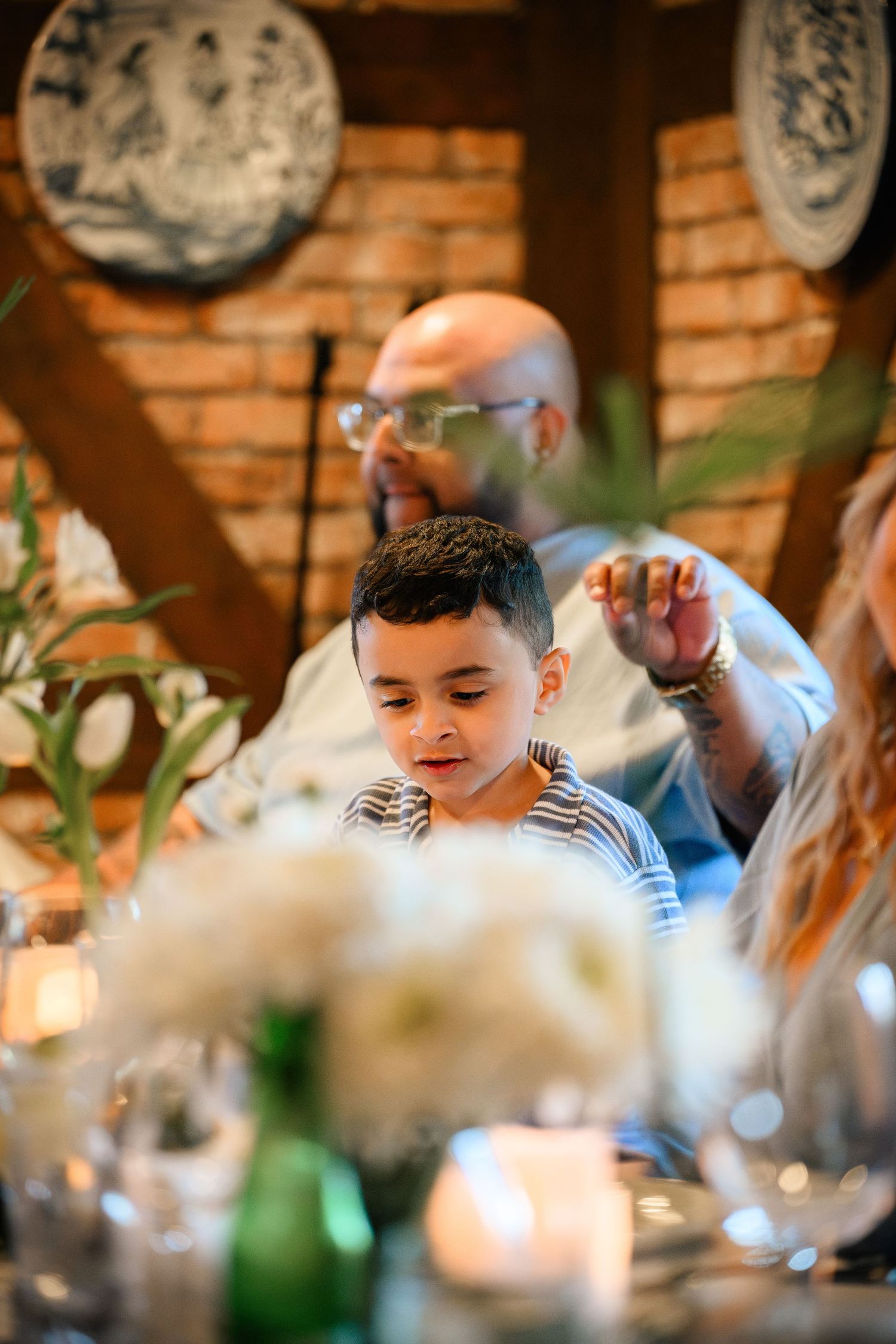 Young boy sitting at an elegant table setting with blurred decorative elements in warm lighting at a restaurant or event.