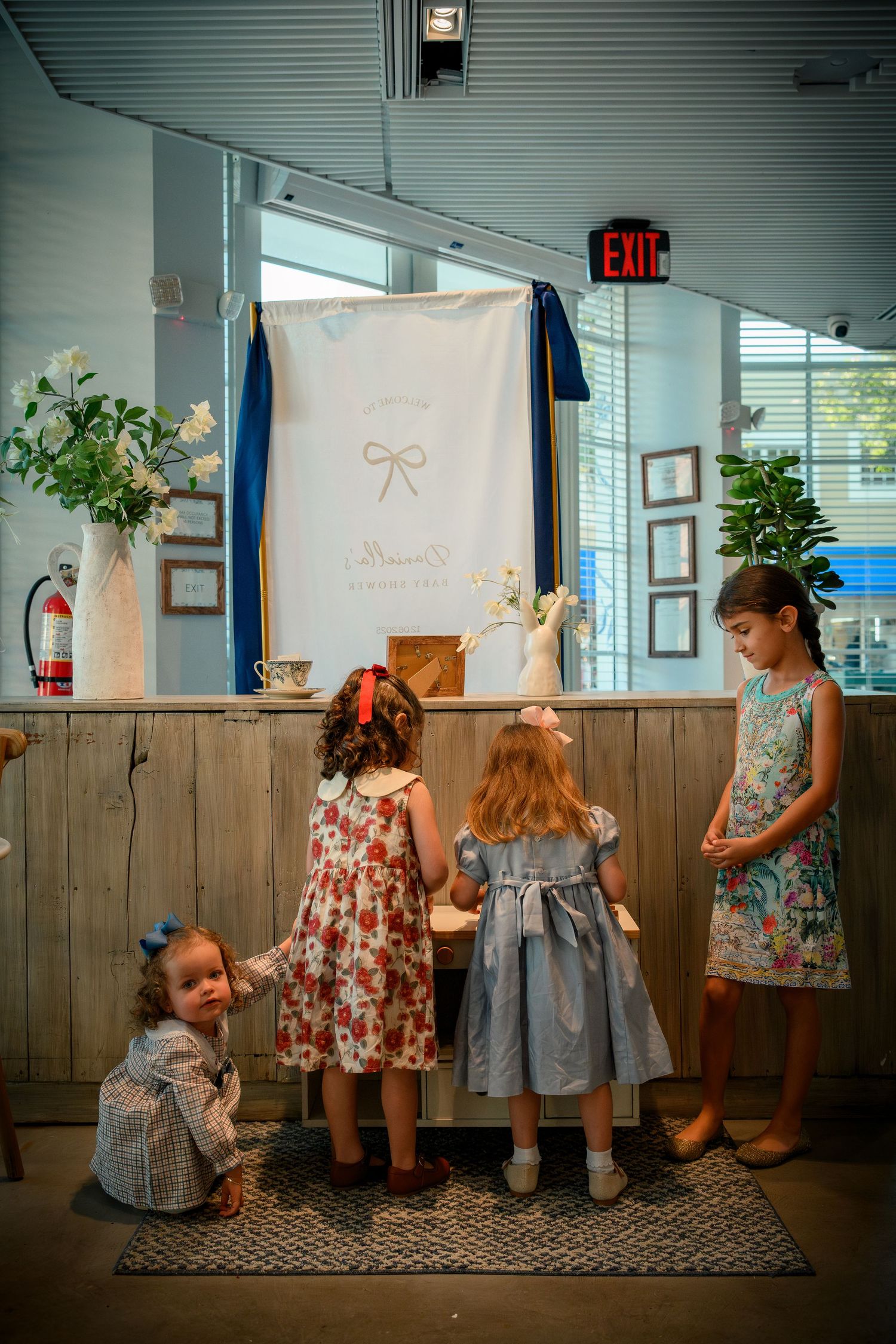 Children in floral and blue dresses stand against wooden paneled wall in an indoor setting with an exit sign visible.