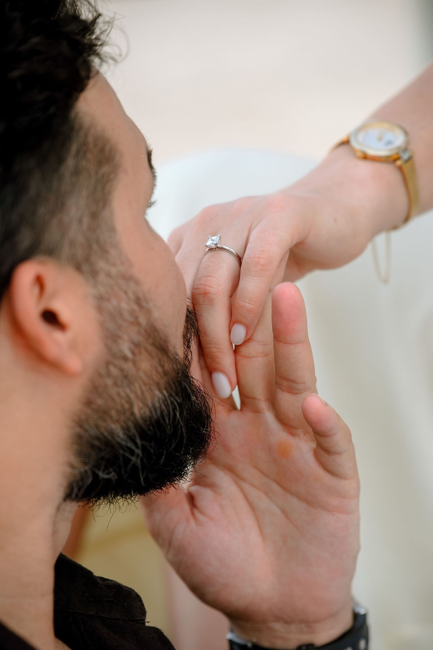 A woman with a diamond ring and gold watch touches a bearded man's face during a grooming session.