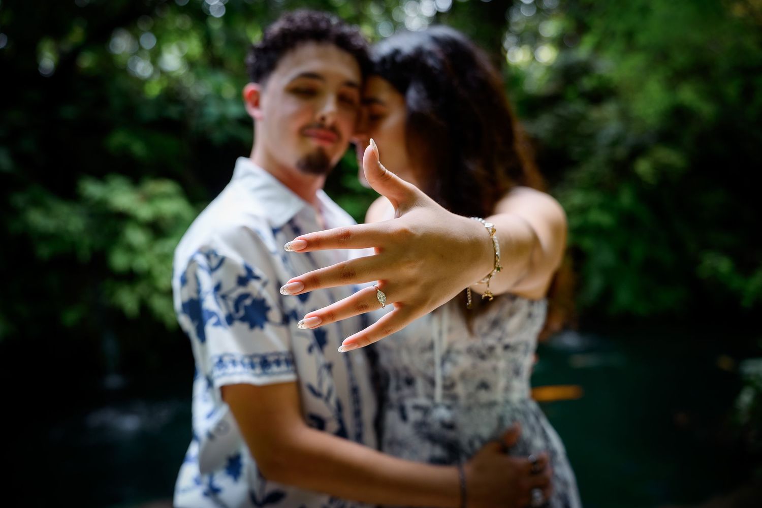 Couple embracing outdoors, woman showing engagement ring on her hand, lush green trees in background.