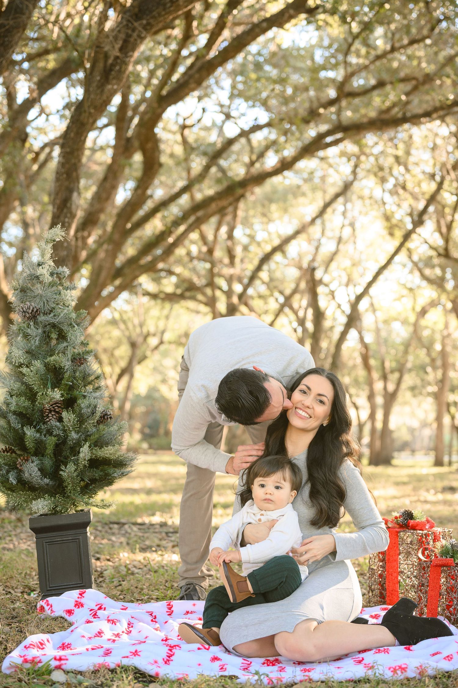 Family Christmas photo with parents and baby on blanket under oak trees outdoors.