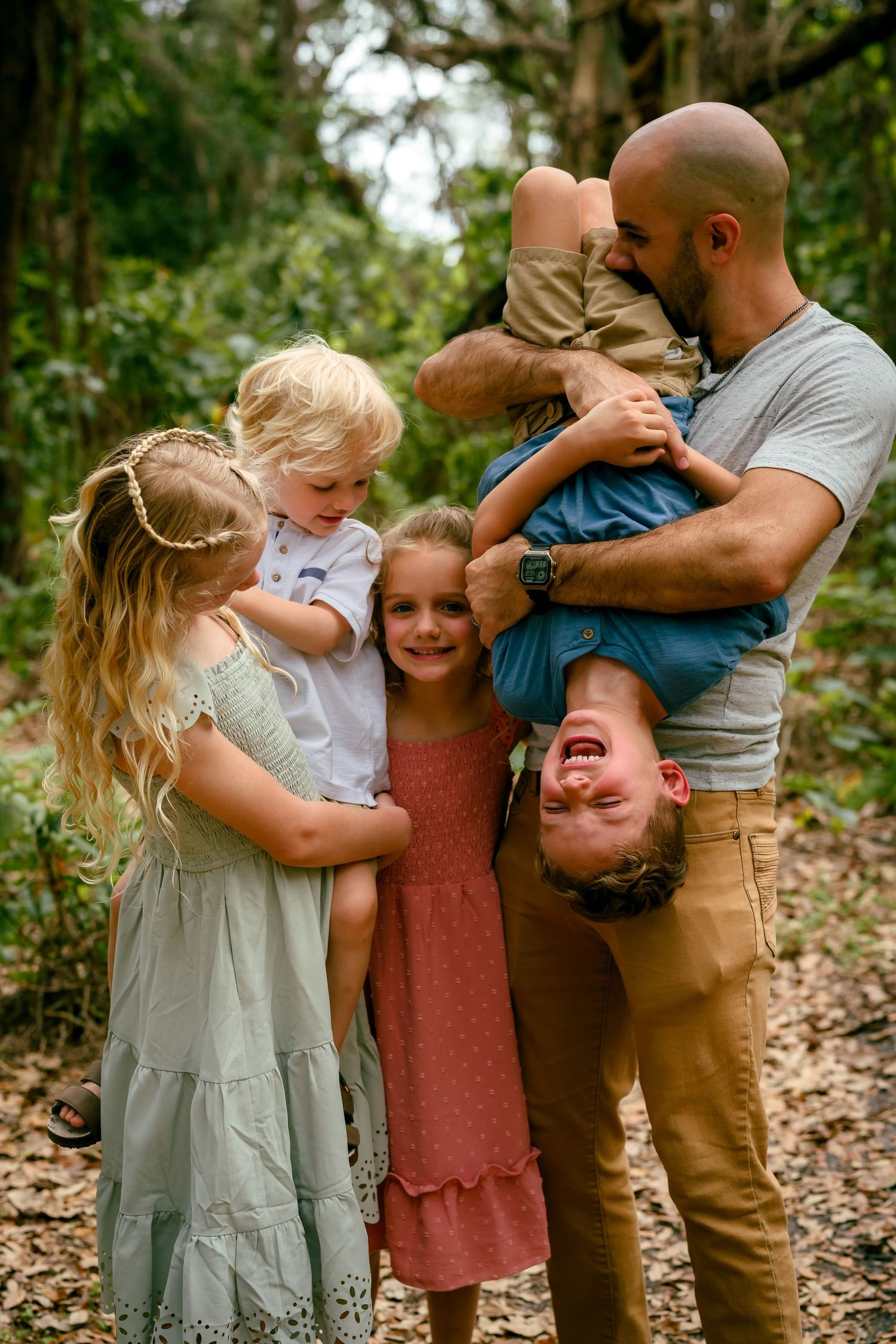 A joyful family of six poses outdoors in a forest, with a bald dad playfully holding a laughing child upside down.