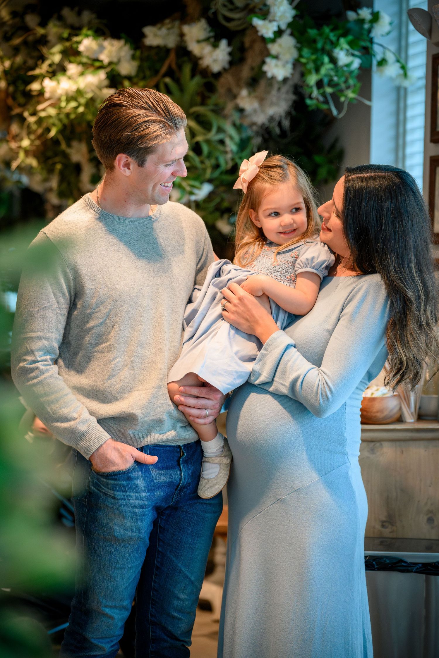 Expectant parents share a tender moment with their young child in a cozy indoor setting with greenery in the background.