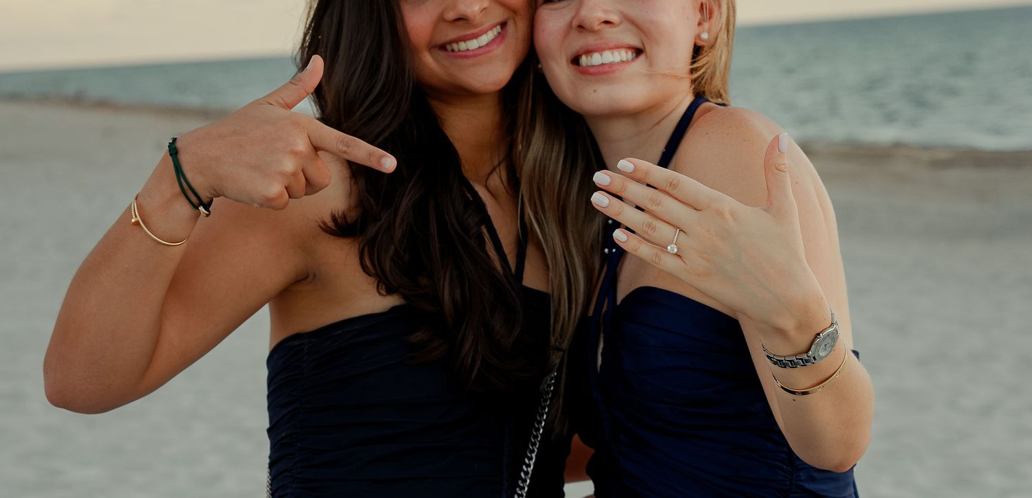 Two smiling women in navy dresses pose together on a beach, showing off bracelets and rings.