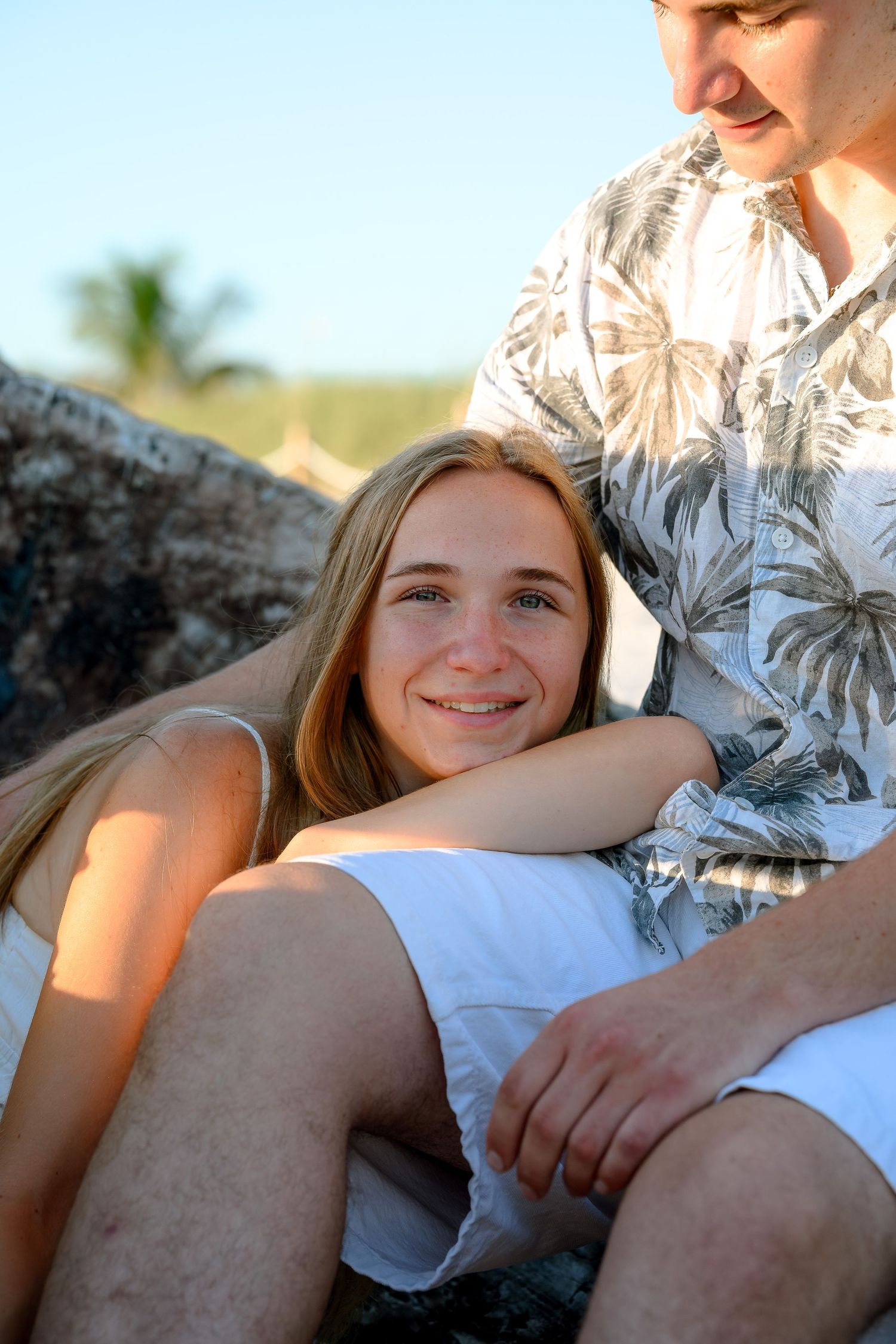 Smiling young blonde woman relaxing outdoors with a man in a floral shirt, sitting on rocks in a sunny tropical setting.