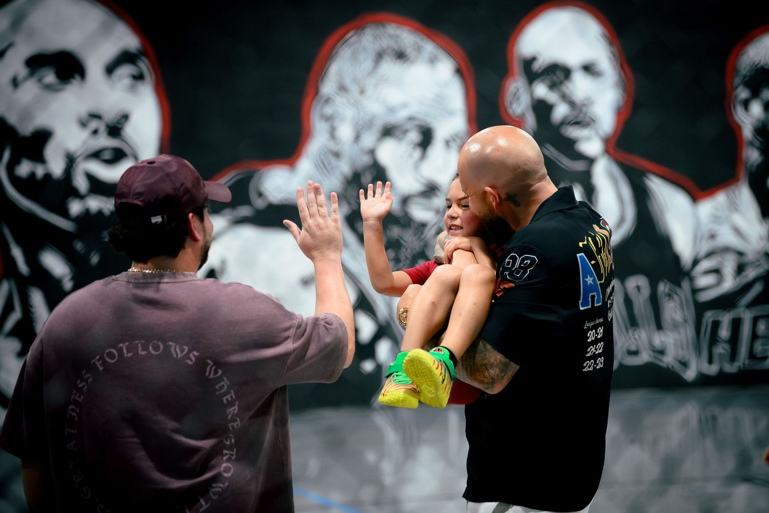 Man holds child up for high-five in front of large mural featuring famous figures at outdoor event.