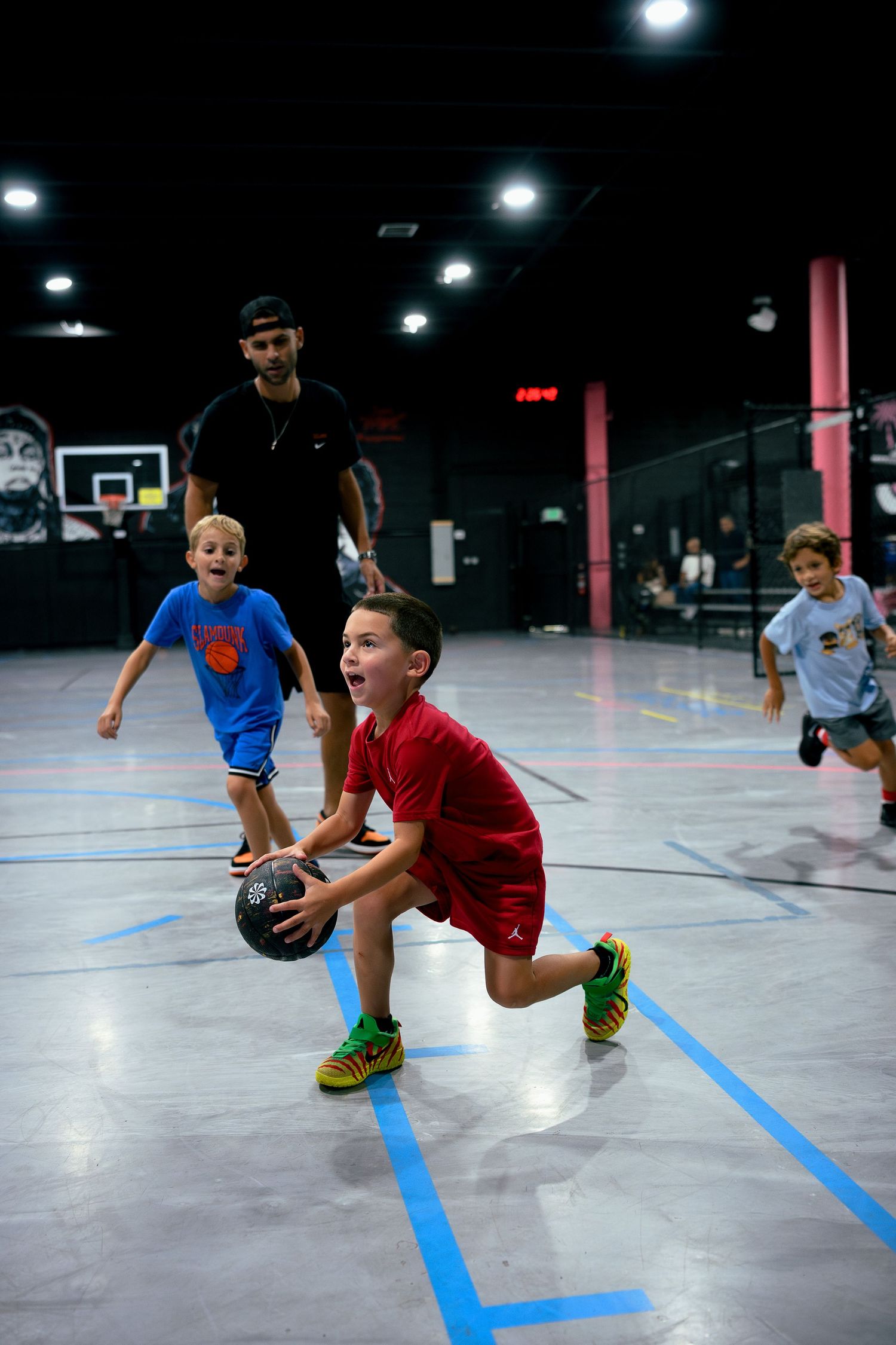 Young boy in red jersey dribbling basketball at indoor court during kids basketball camp with coach watching.