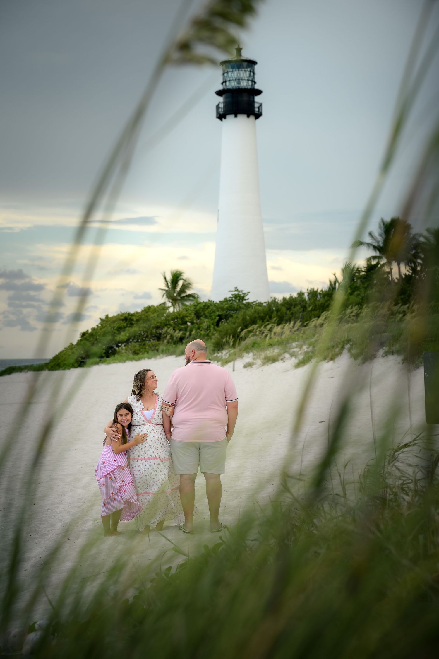 Family of three standing on sandy beach path near Cape Florida Lighthouse at dusk.