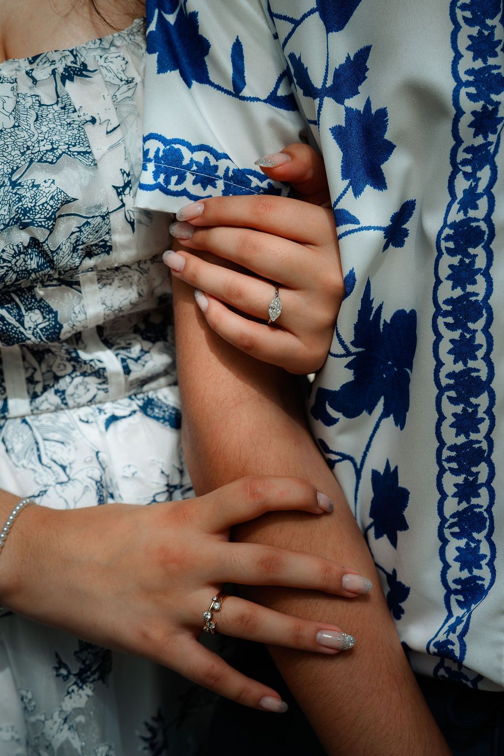 Two hands with elegant rings and manicured nails clasped together, wearing blue floral embroidered garments.