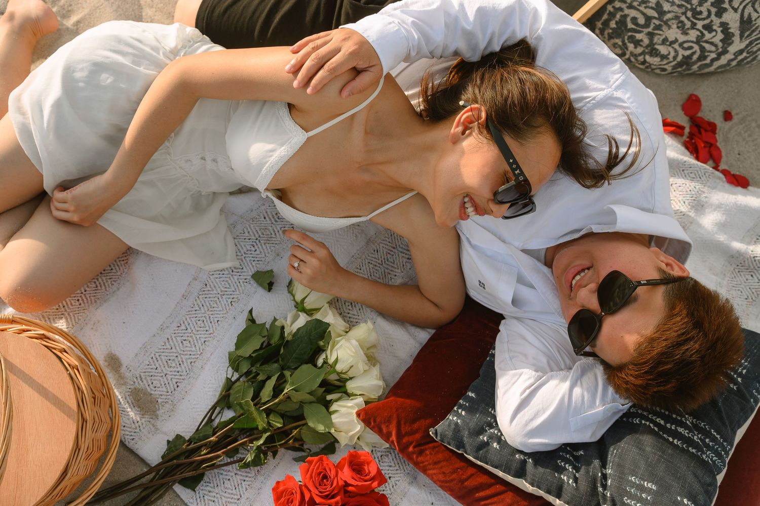 Romantic couple lying together on a blanket surrounded by red and white roses on a sunny day.