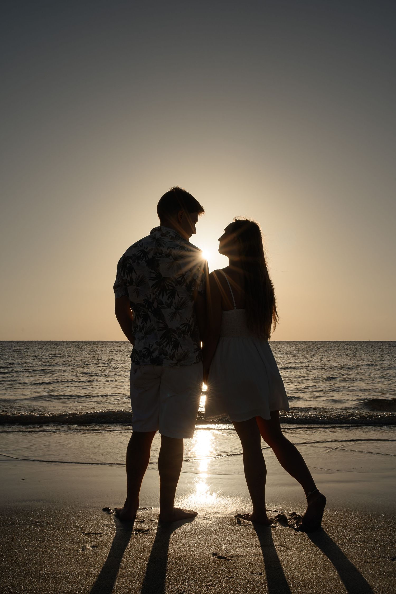 Romantic couple silhouetted against a golden sunset on a beach, standing close together near the ocean waves.
