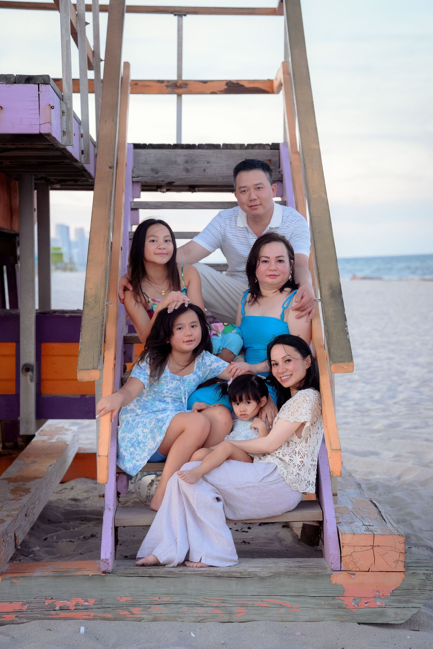 Asian family of five posing together on beach stairs near a lifeguard tower at dusk.