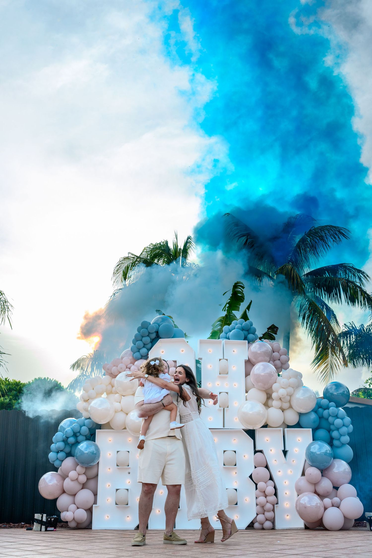 A group poses together in white outfits with pink and blue balloon decorations and smoke effects in an outdoor setting.