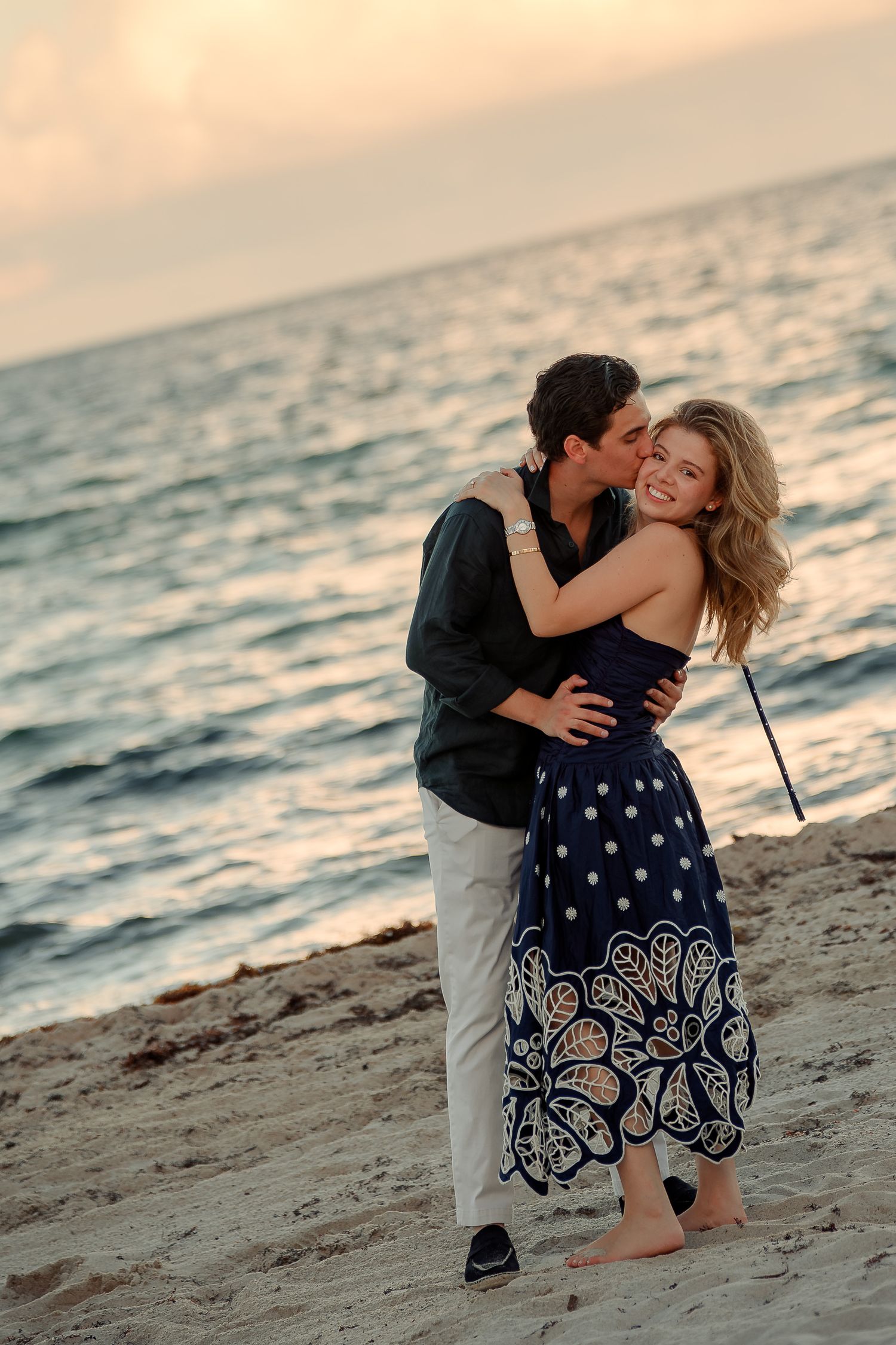 Couple sharing a romantic kiss on a sandy beach at sunset, woman in blue floral dress smiling joyfully.