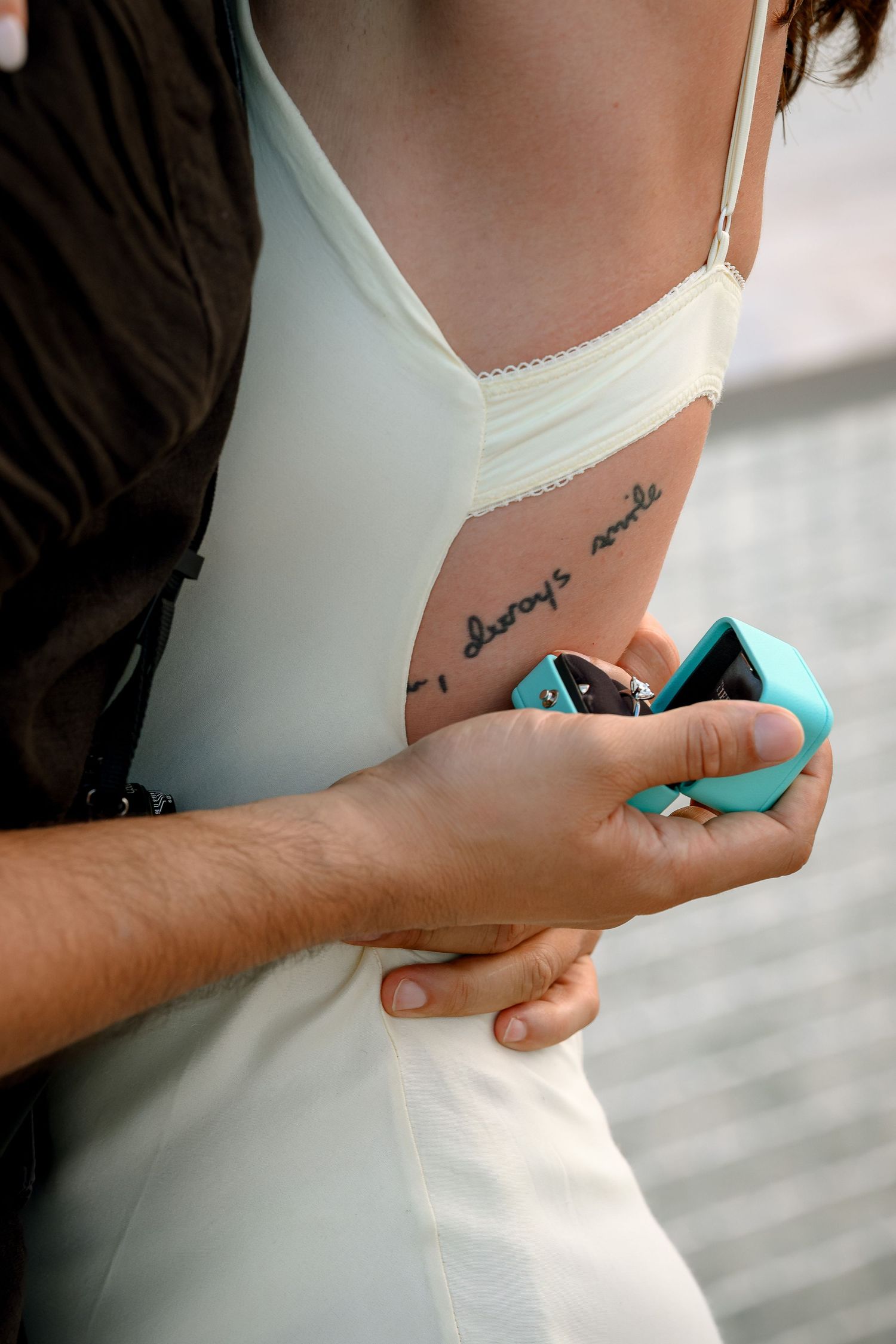Close-up of a couple embracing, woman in white dress with cursive tattoo, man holding teal phone.