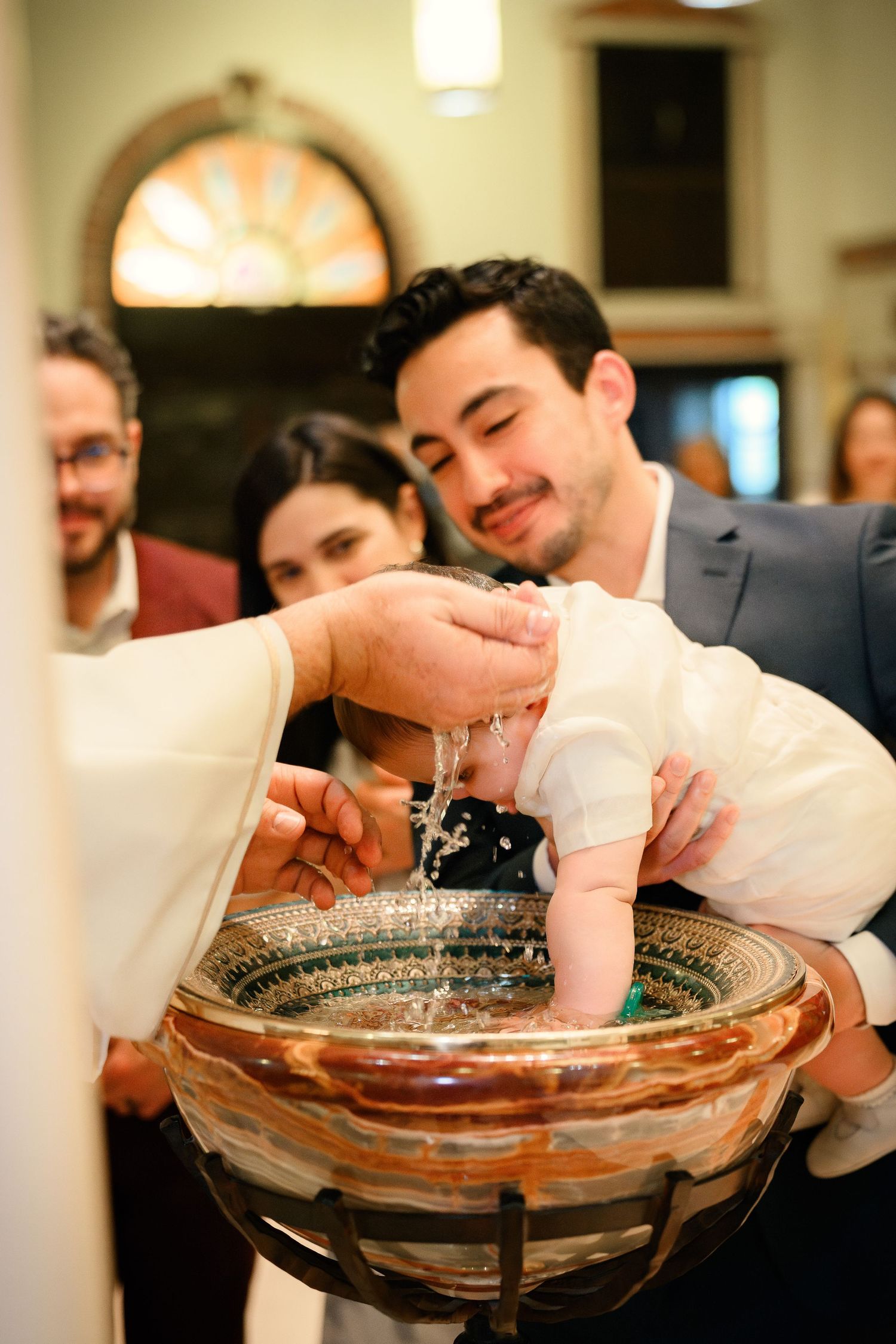 A man holds a baby during a baptism ceremony with water being poured over the infant.