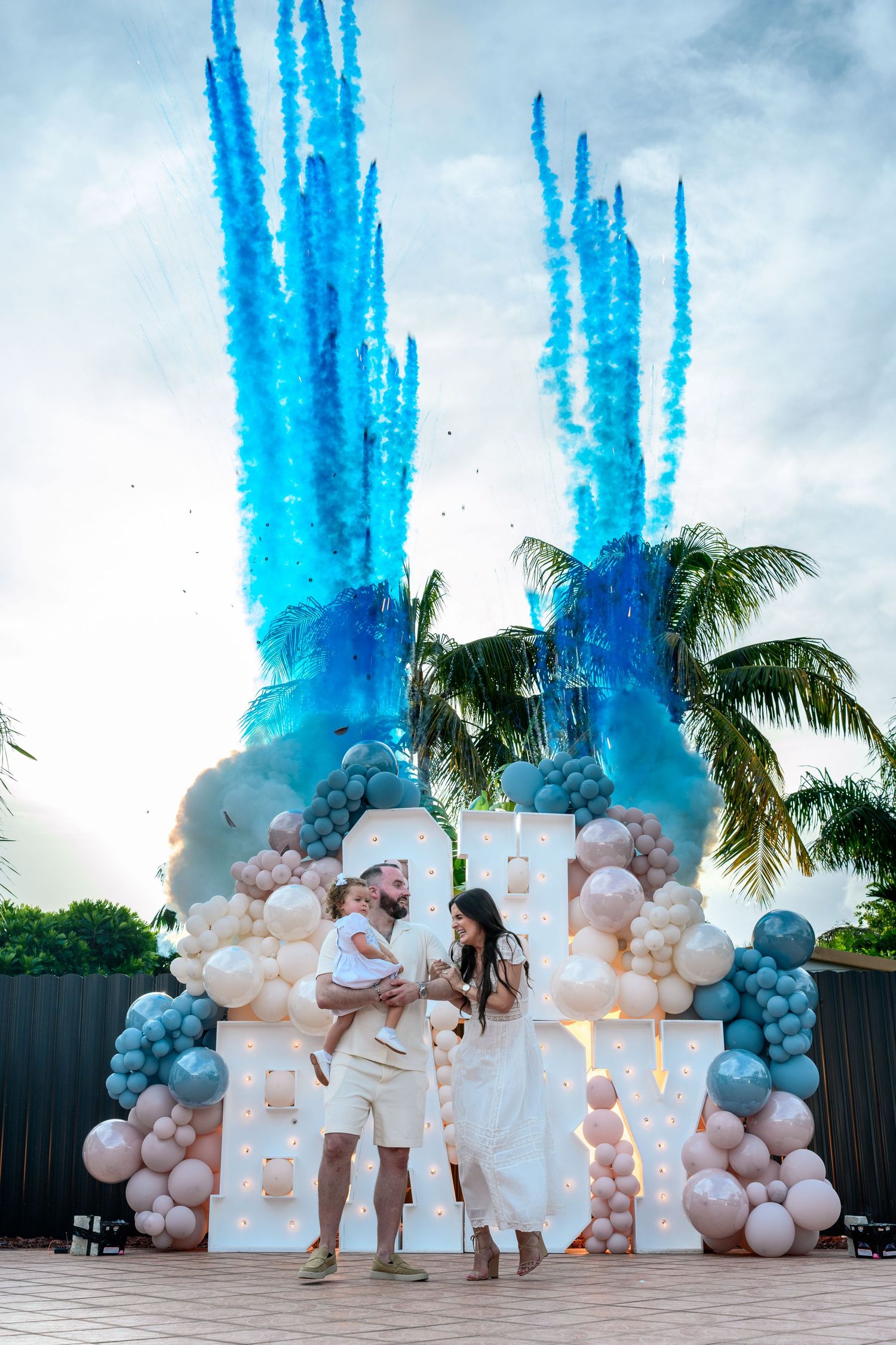 Family gender reveal party with blue smoke cannons, balloon arch decor, and palm trees in outdoor celebration.