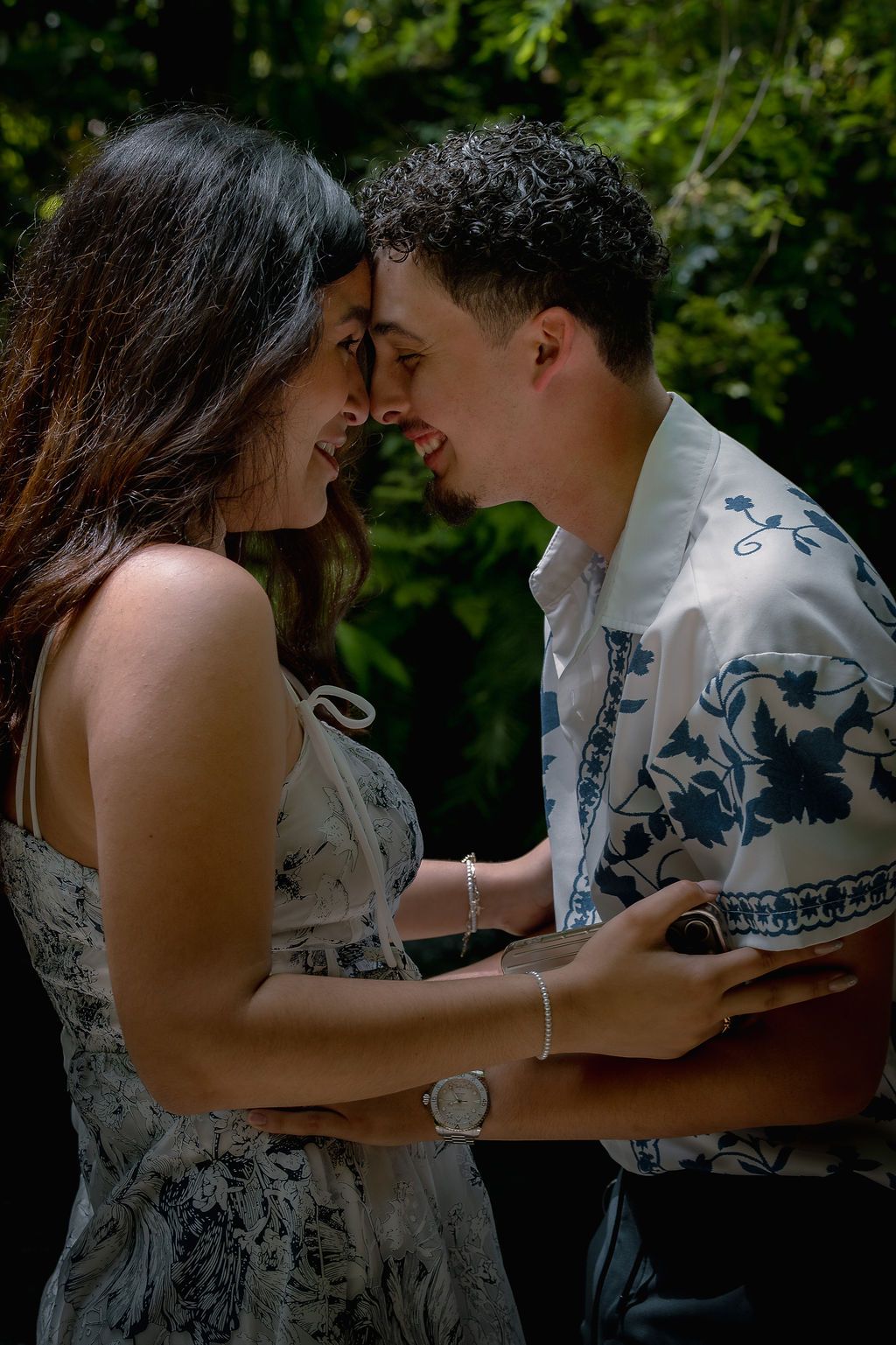 A young couple shares a romantic moment outdoors, smiling and touching foreheads amid lush green foliage.