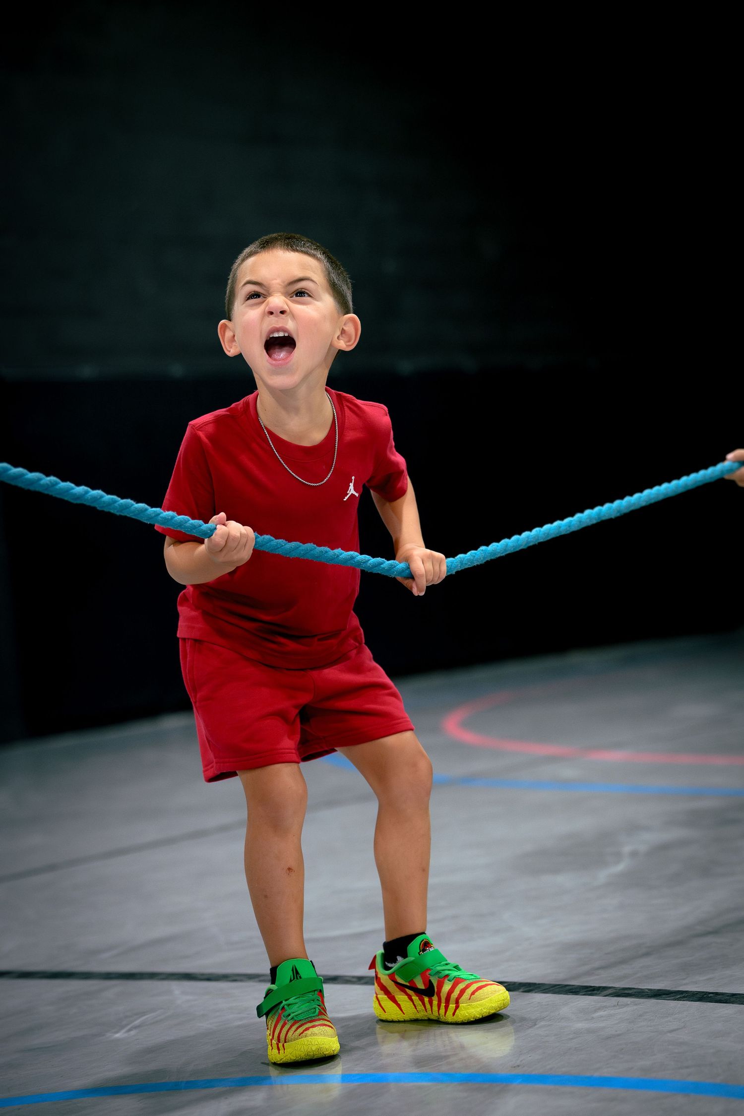 Young boy in red Jordan outfit pulling a blue rope with mouth open, competing in a gym tug-of-war activity.
