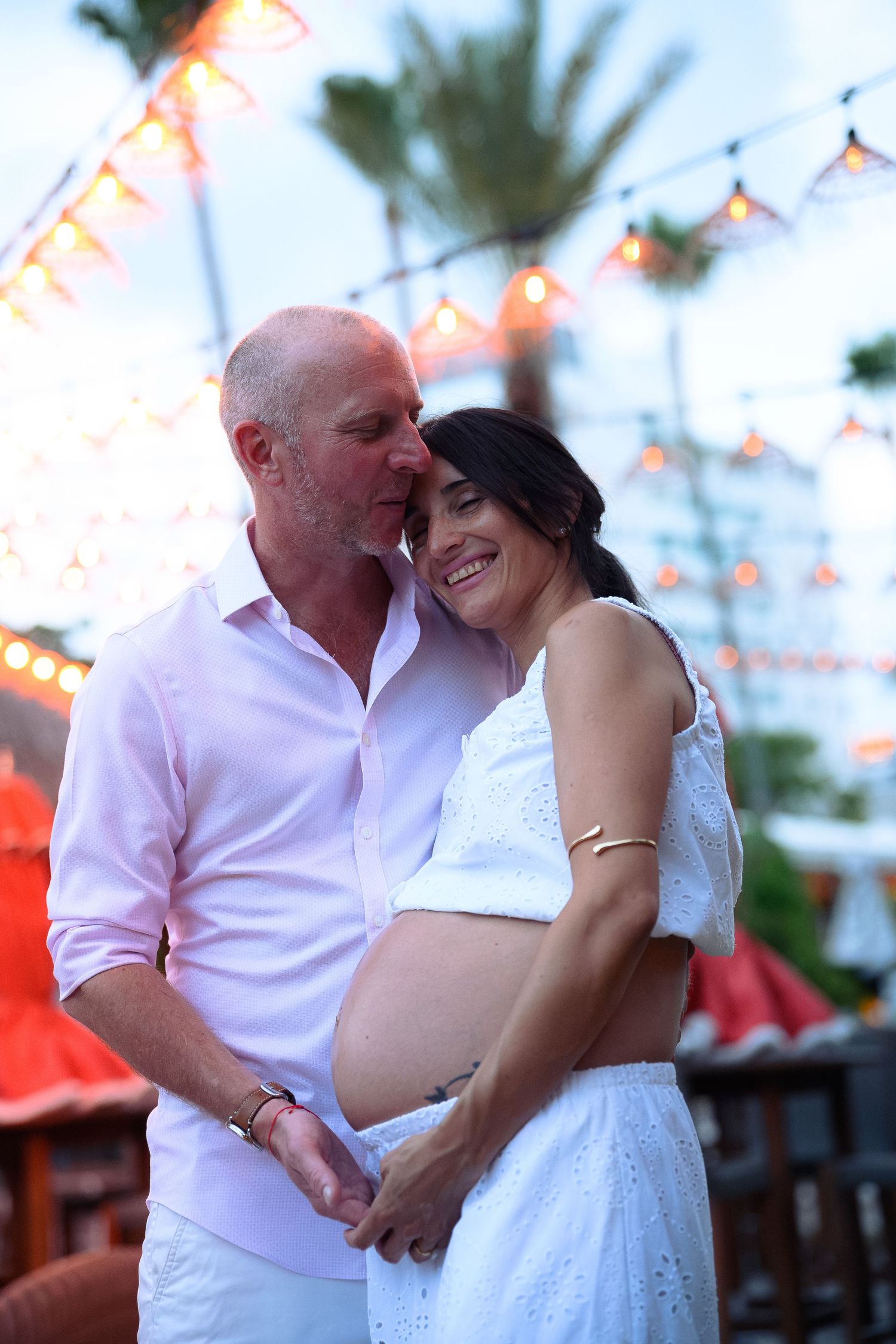Happy couple in white outfits sharing a tender maternity moment outdoors with string lights and palm trees.