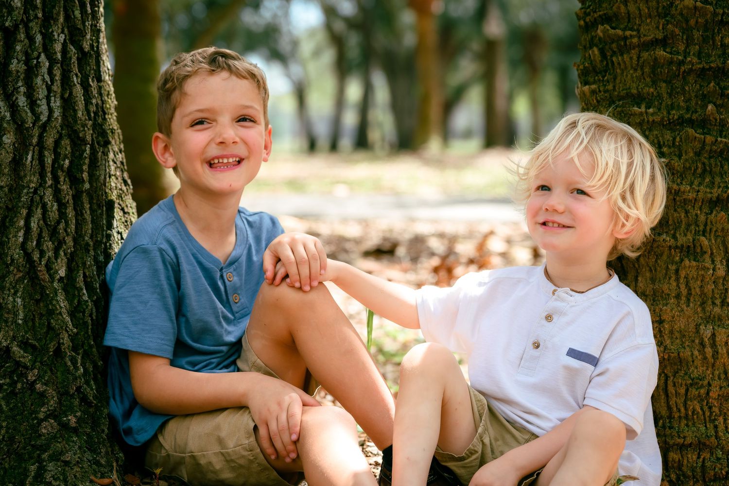 Two smiling young boys sitting together against a tree trunk in a sunny park setting.