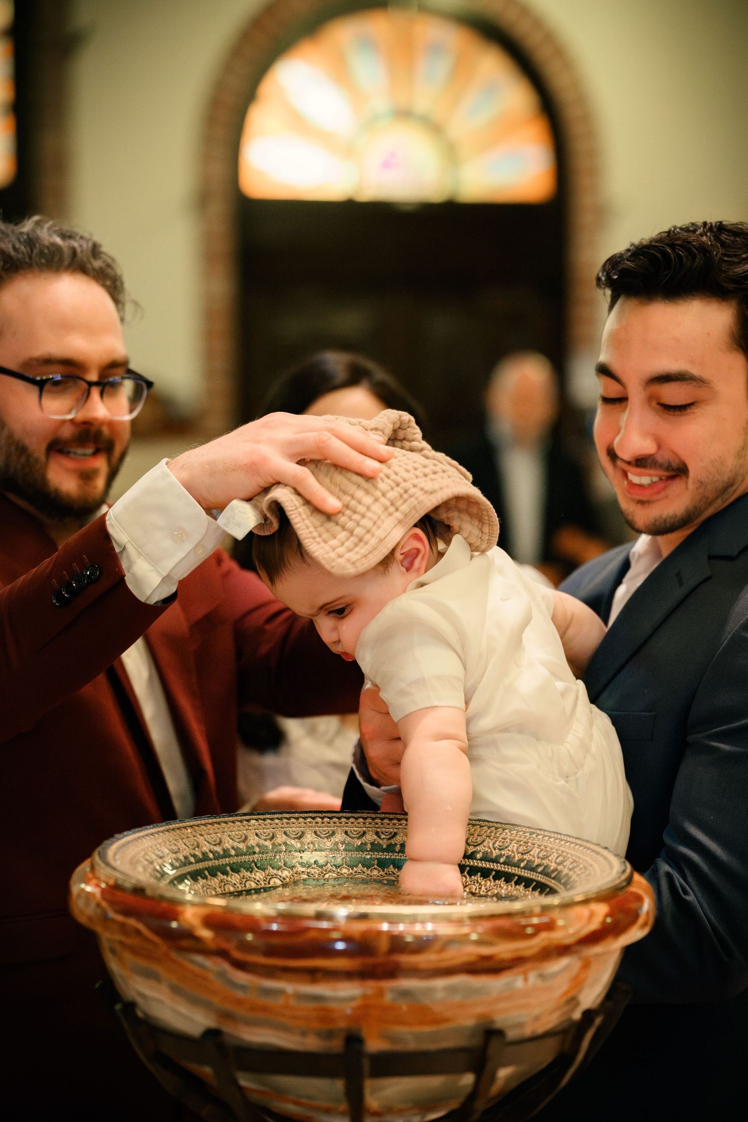 A baby in white clothing is being baptized in a glass baptismal font during a religious ceremony.