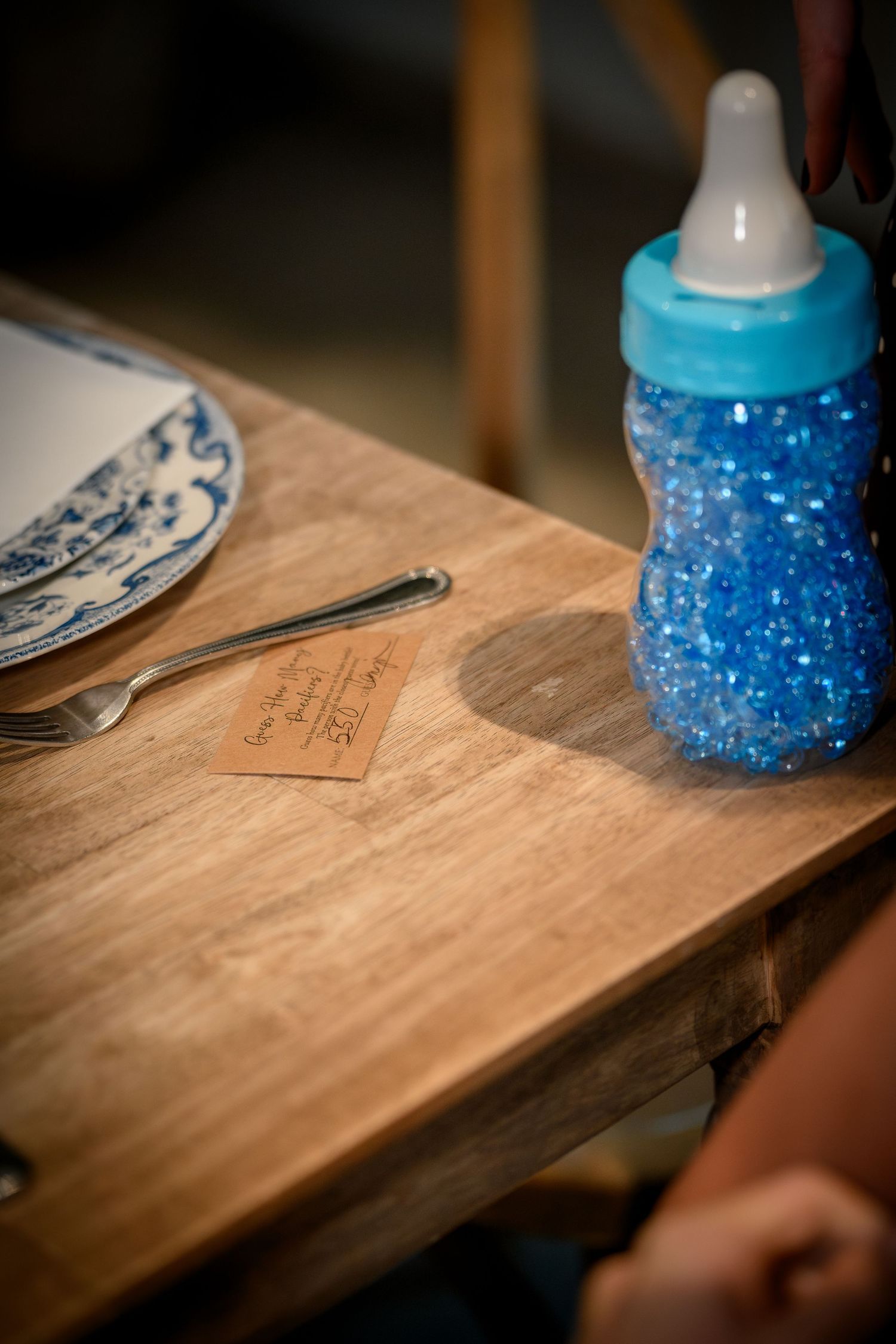 A blue baby bottle sits on a rustic wooden table with a place setting visible in the background.