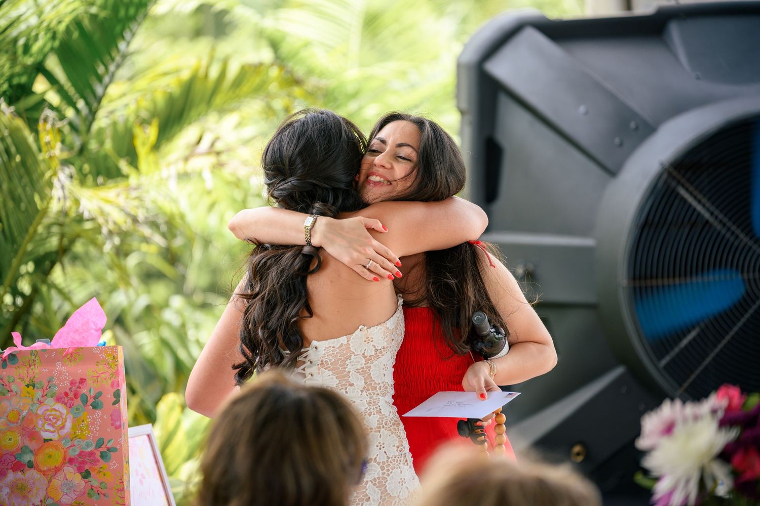 Two people share a warm embrace at an outdoor event with lush greenery in the background.