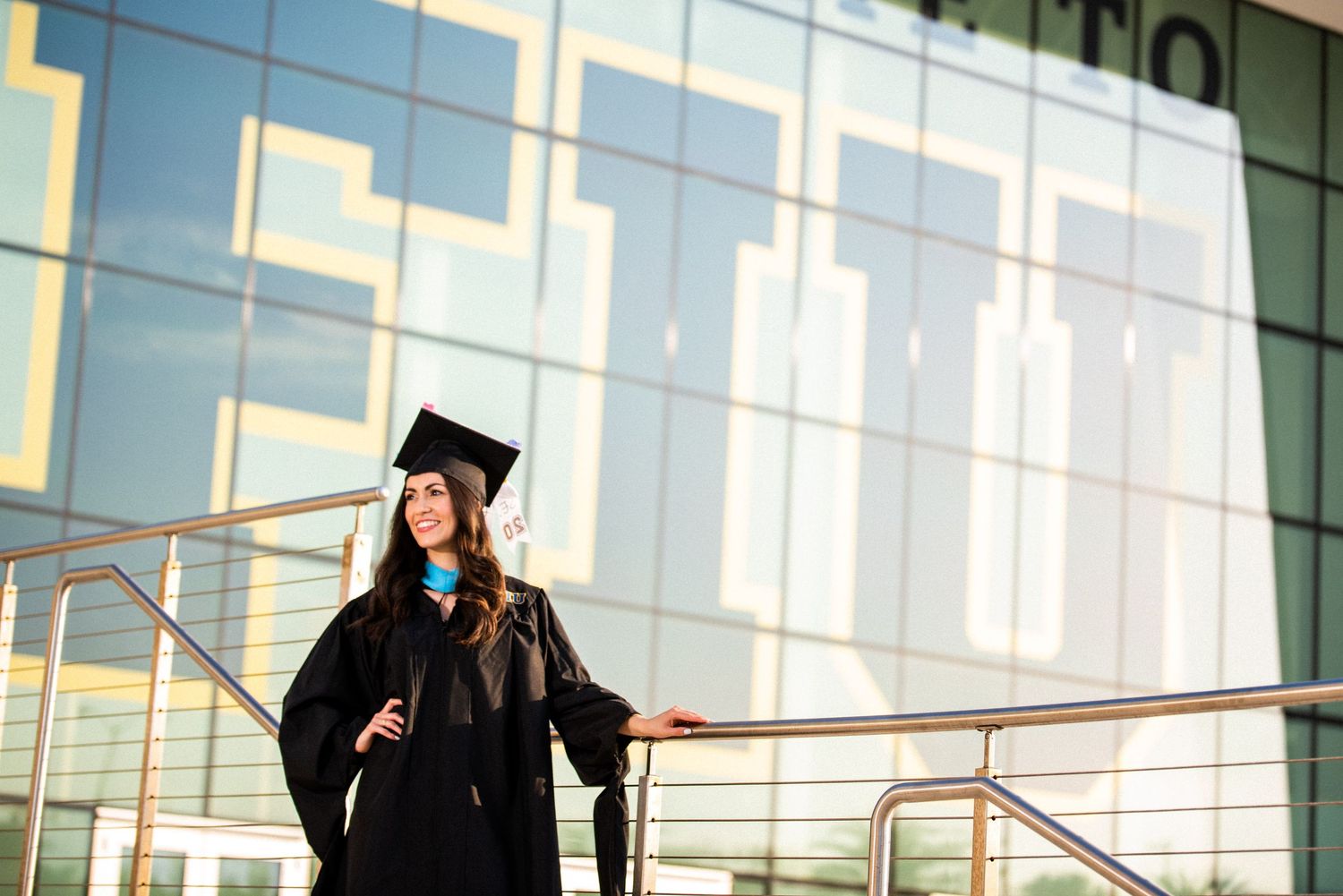 Female graduate in black cap and gown posing confidently outside a modern glass university building.