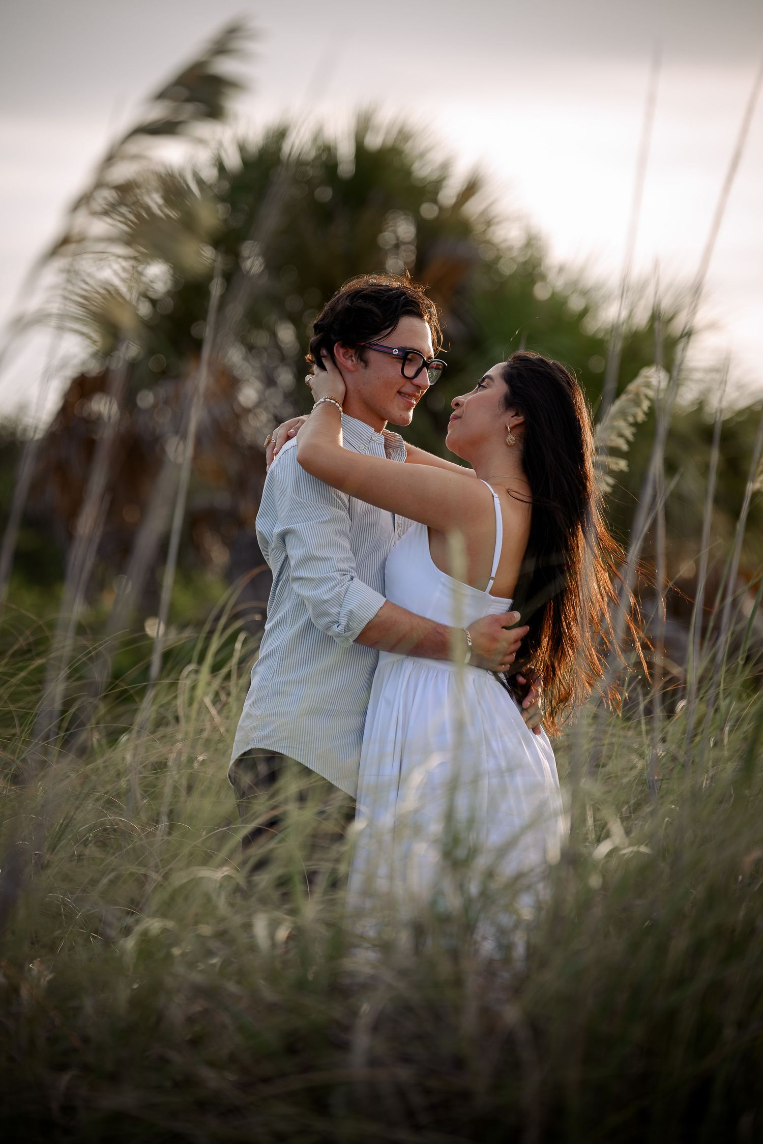 A couple embraces romantically in tall coastal grass, woman in white dress, man in glasses, at golden hour.