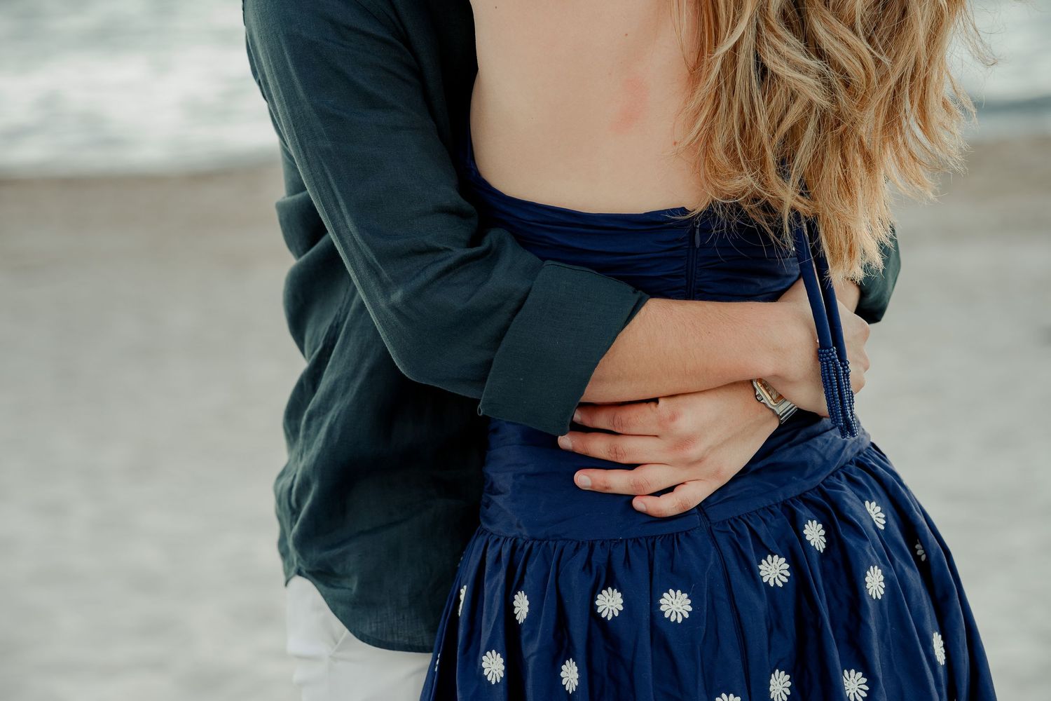 Couple embracing on a beach, woman in blue floral dress, man in dark jacket, romantic outdoor portrait.