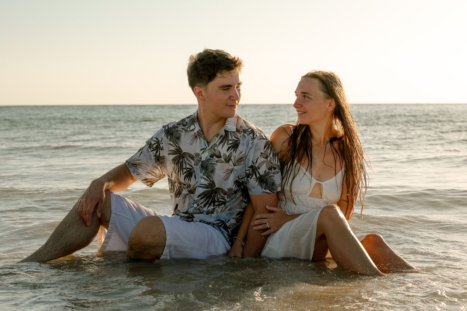 Couple sitting together in shallow ocean water at sunset, wearing white outfits, smiling at each other on a tropical beach.